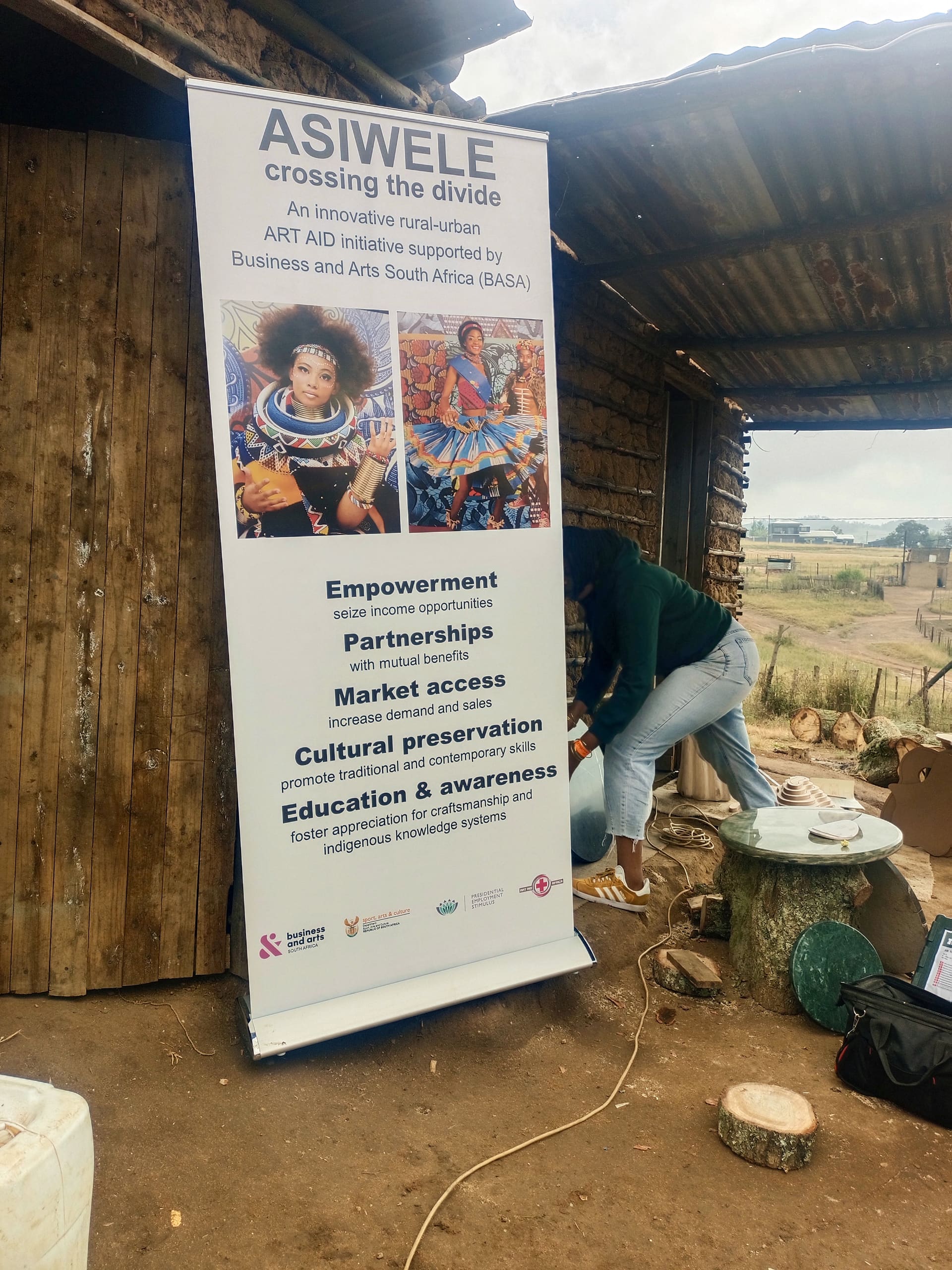 A person adjusts a tall banner beside a rustic wooden building. The banner, titled 