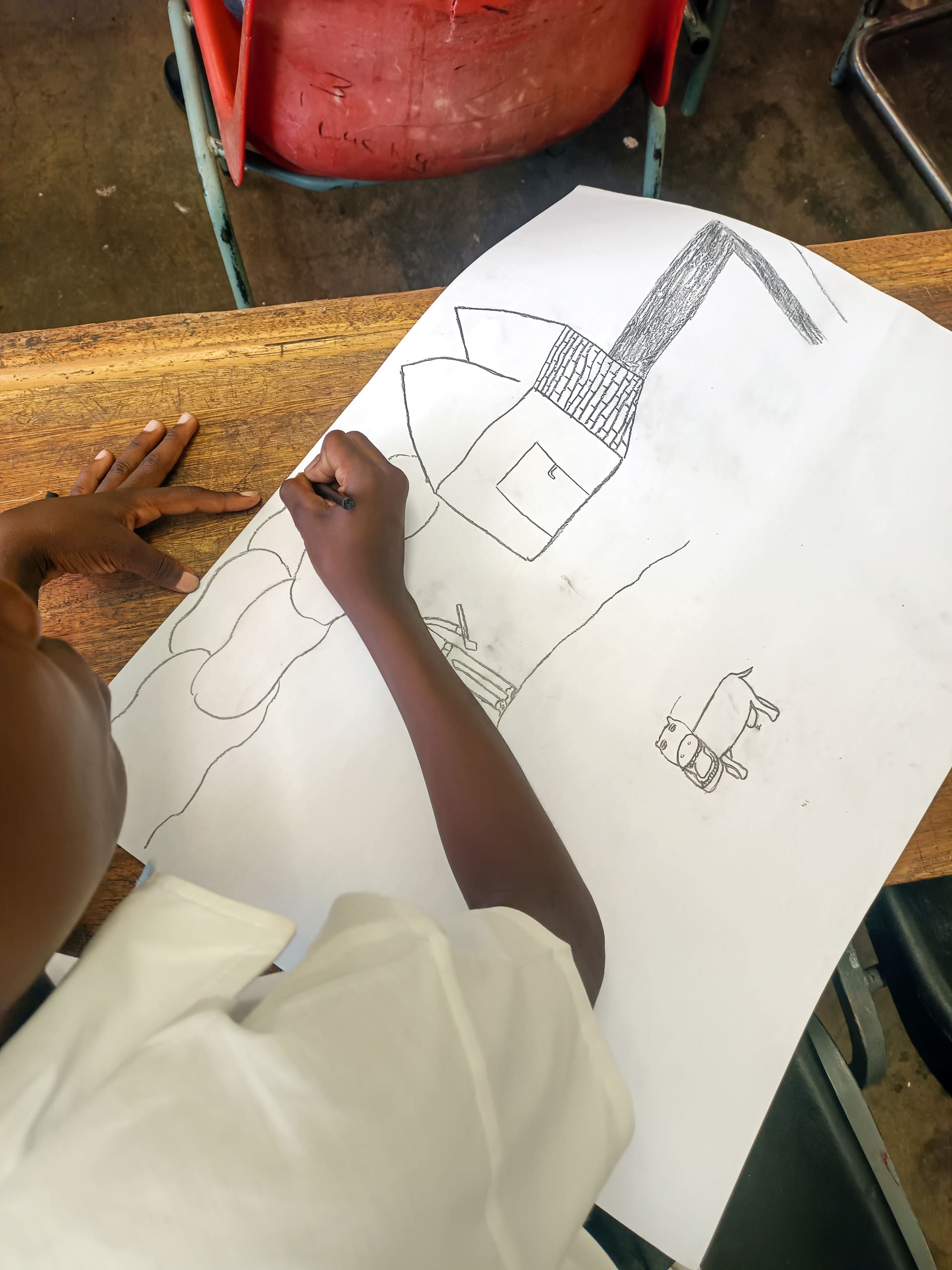 A child in a white shirt draws with a pencil on a large sheet of paper at a wooden desk. The drawing features shapes resembling a house, a tree, and a car. The child’s left hand holds the paper steady whilst they draw with their right hand.