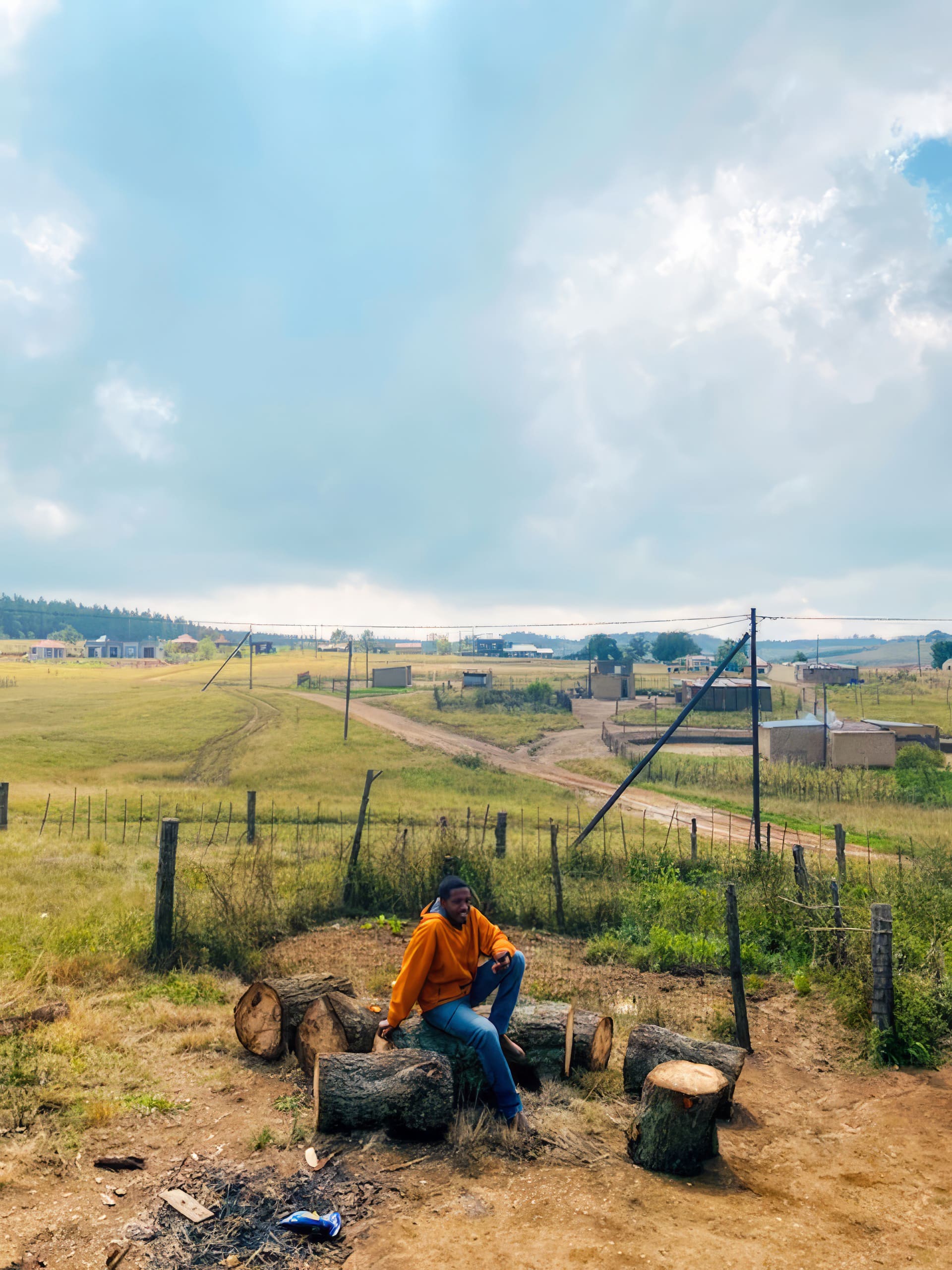 A person in an orange jumper sits on a log among cut tree stumps in a rural, grassy landscape. Dirt roads, power lines, fences, and scattered houses stretch across the background under a partly cloudy sky.