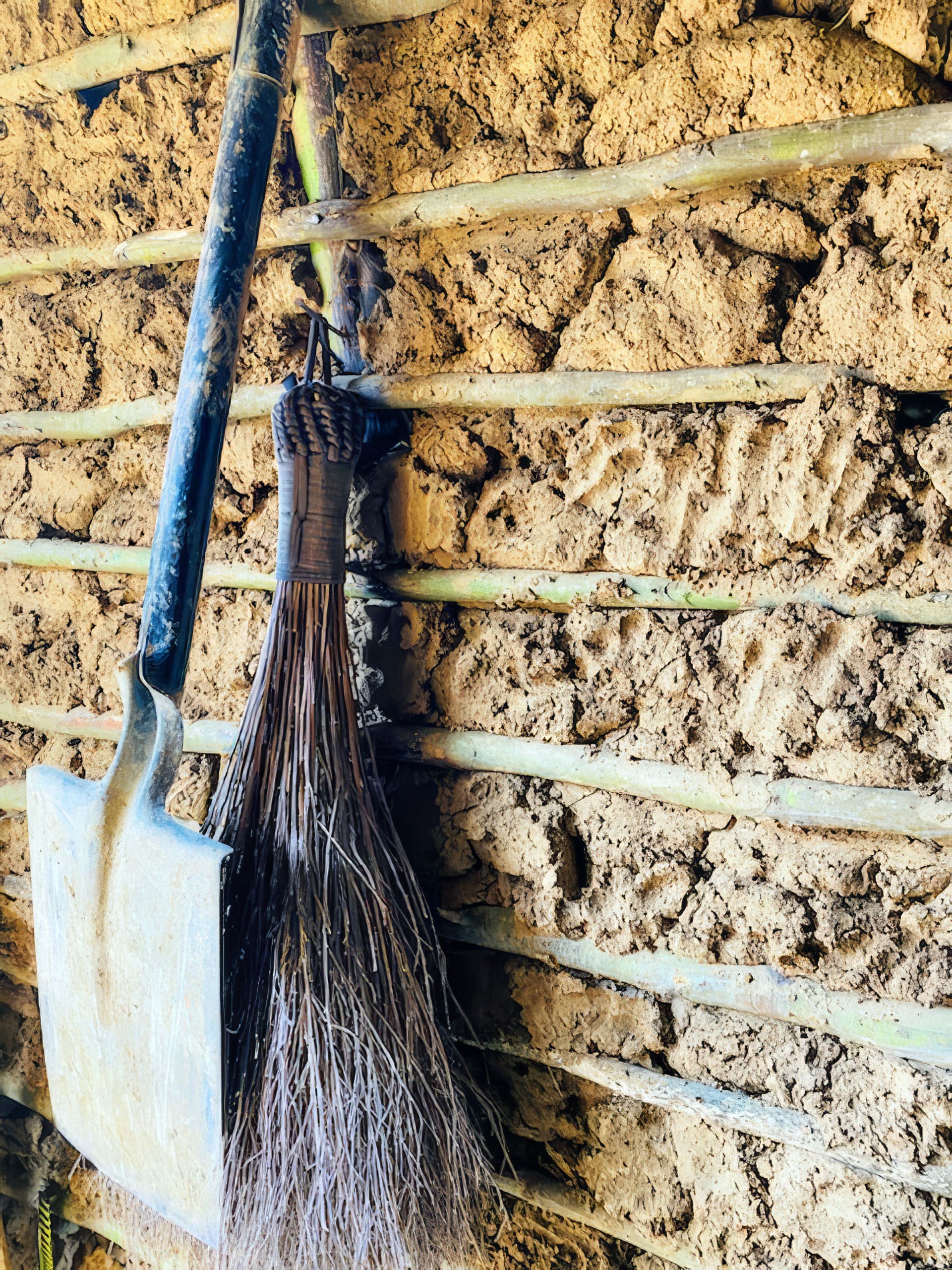 A metal spade and a traditional broom made of twigs hang on a wall made of mud and bamboo canes. Sunlight softly illuminates the rough texture of the natural materials, giving a rustic, earthy feel to the scene.