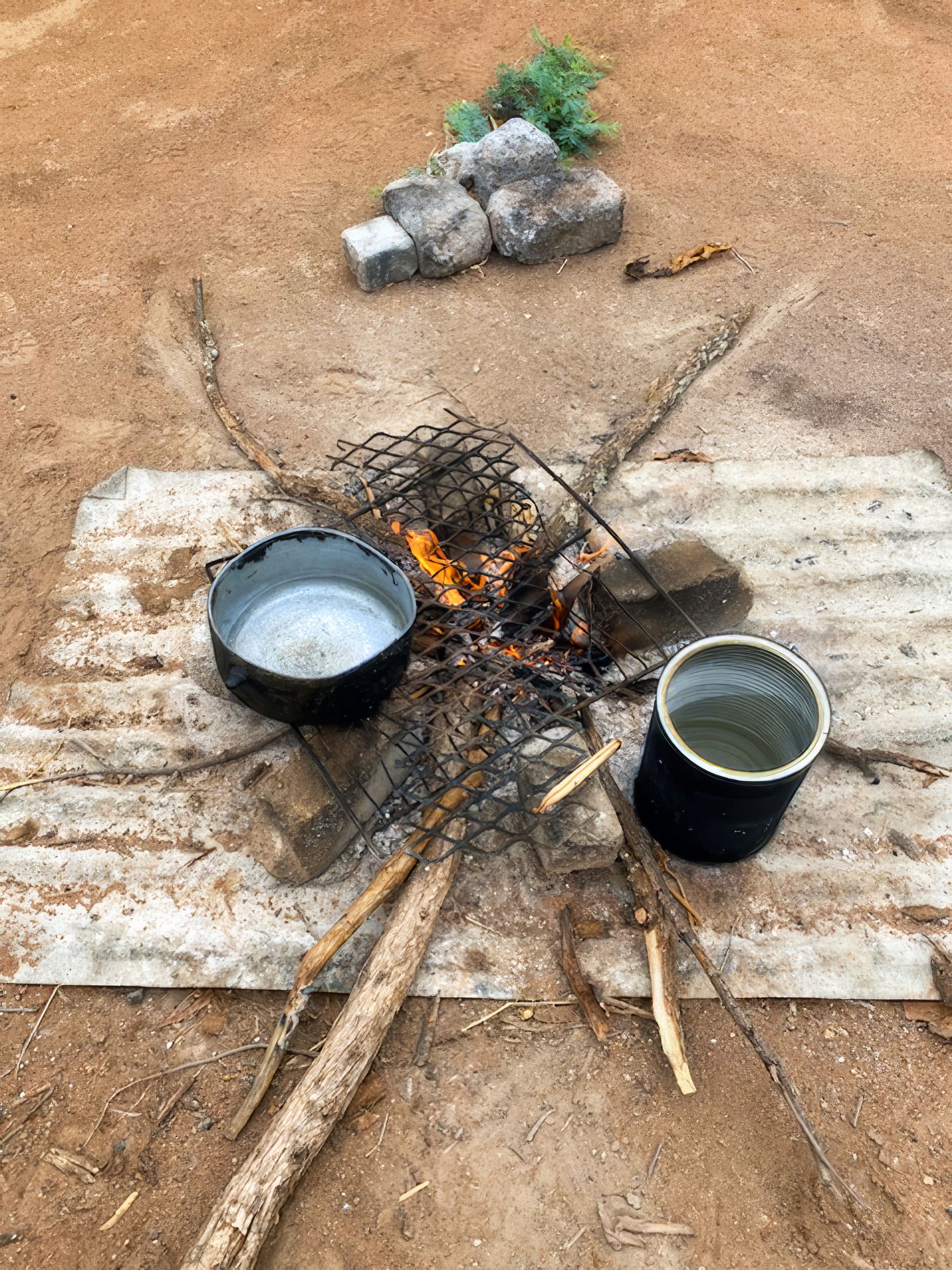 A campfire burns on sandy ground with metal grates arranged over it. A black pot and a metal container sit on the grates. Several sticks support the setup. In the background, stones and some greenery are placed together on the earth.