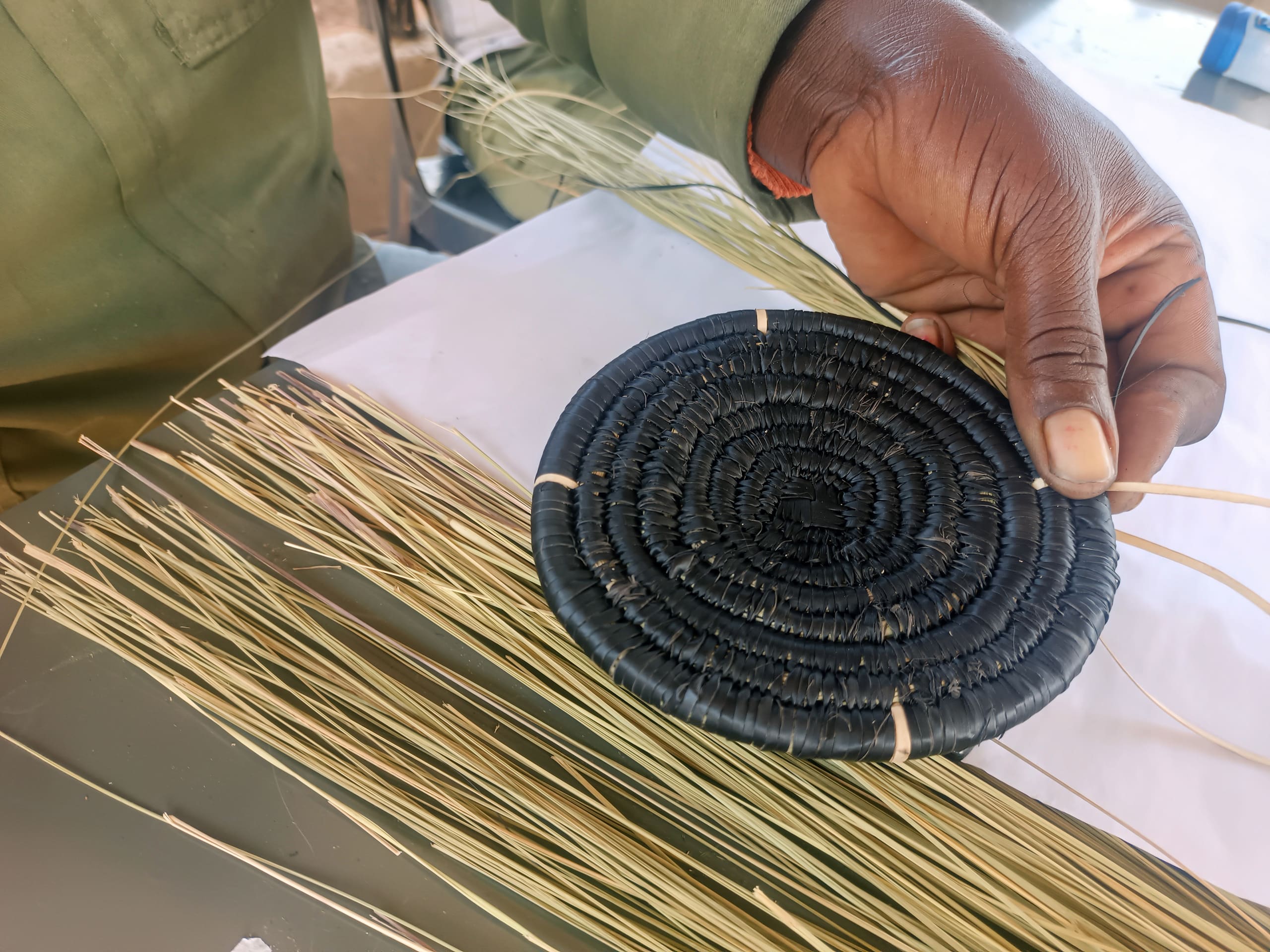 A close-up of a person’s hand weaving a round, black basket from dried grass or reeds. The unfinished basket and weaving materials rest on a table, with the hand guiding thin strands into the circular pattern. The person wears a long-sleeve green shirt.