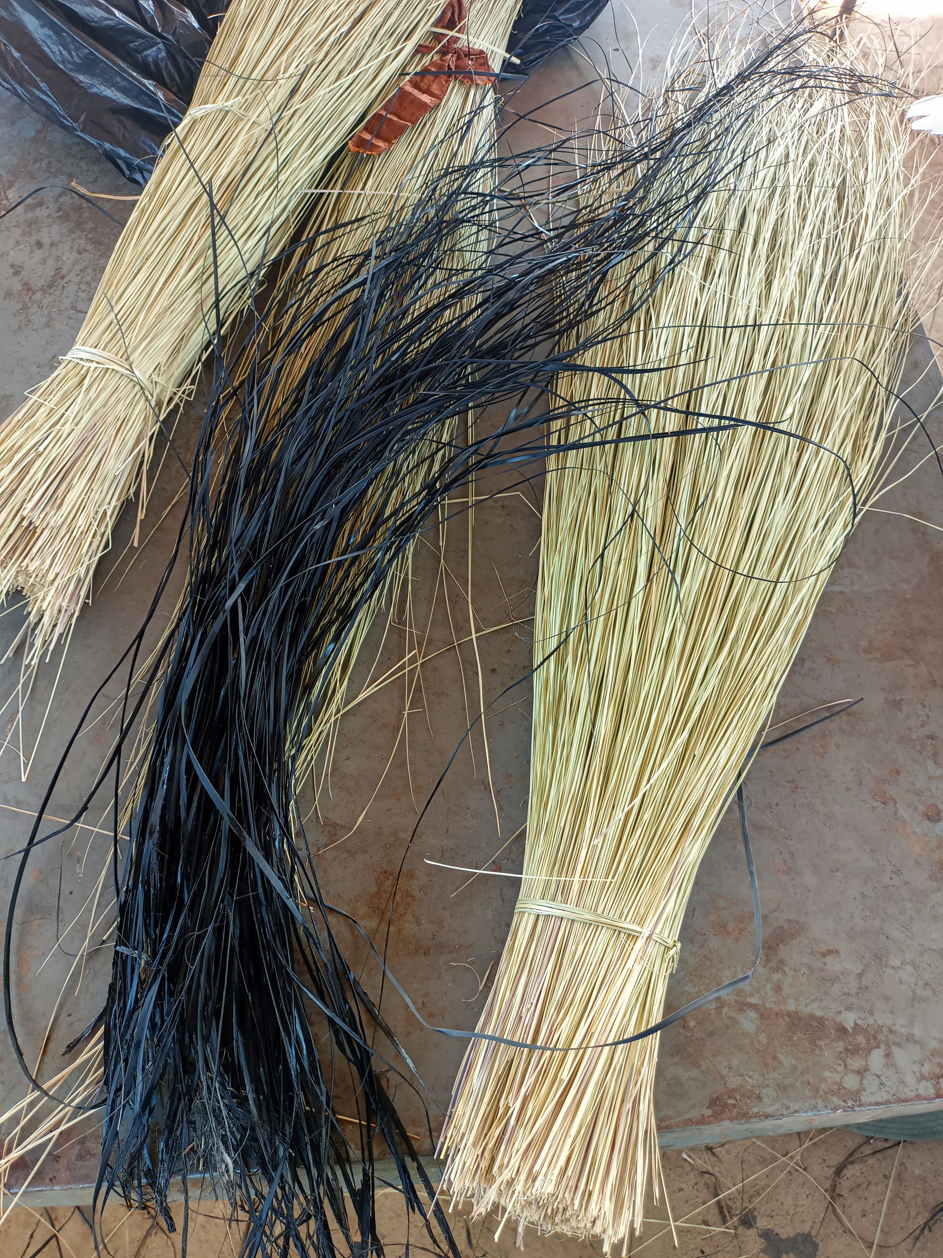 Bundles of long, thin, dried grass or reeds lie on a stone surface. Two light tan bundles are tied with string, while a third bundle is dyed black. The strands are neatly grouped but splay out at the ends, showing varied textures.