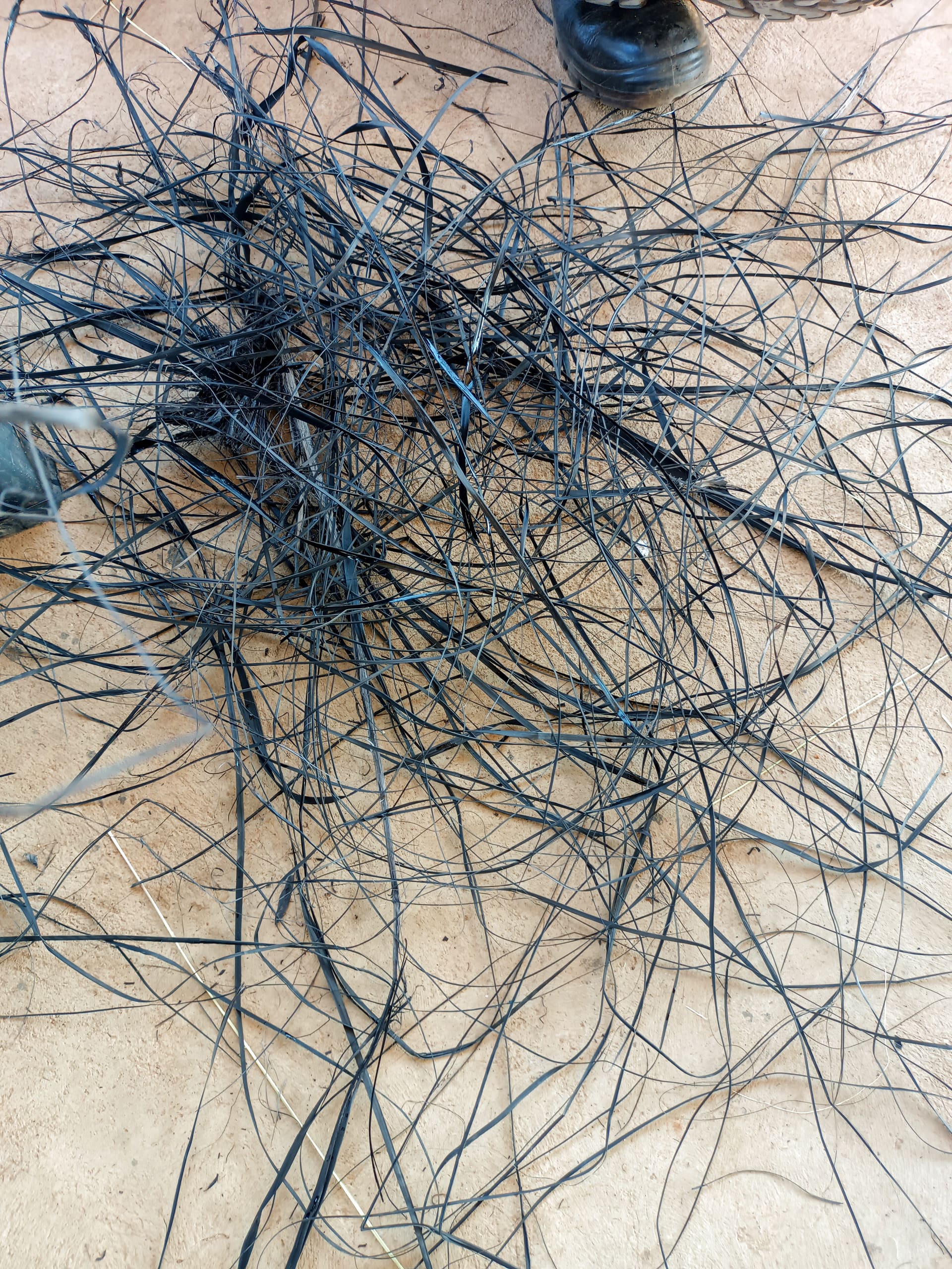 A scattered pile of long, thin, black wires or plastic strips is strewn across a rough, light brown concrete surface. Two black shoes are visible near the edges of the image, framing the tangled pile.