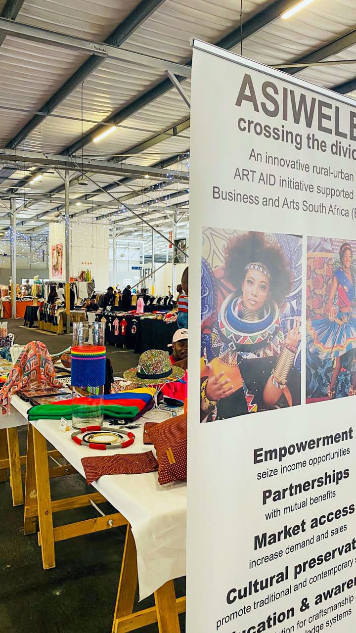 A vibrant indoor market scene with handmade crafts, jewellery, and colourful textiles displayed on tables. A large banner reads “ASIWELE: crossing the divide,” with photos of African women in traditional attire and text about empowerment and partnerships.