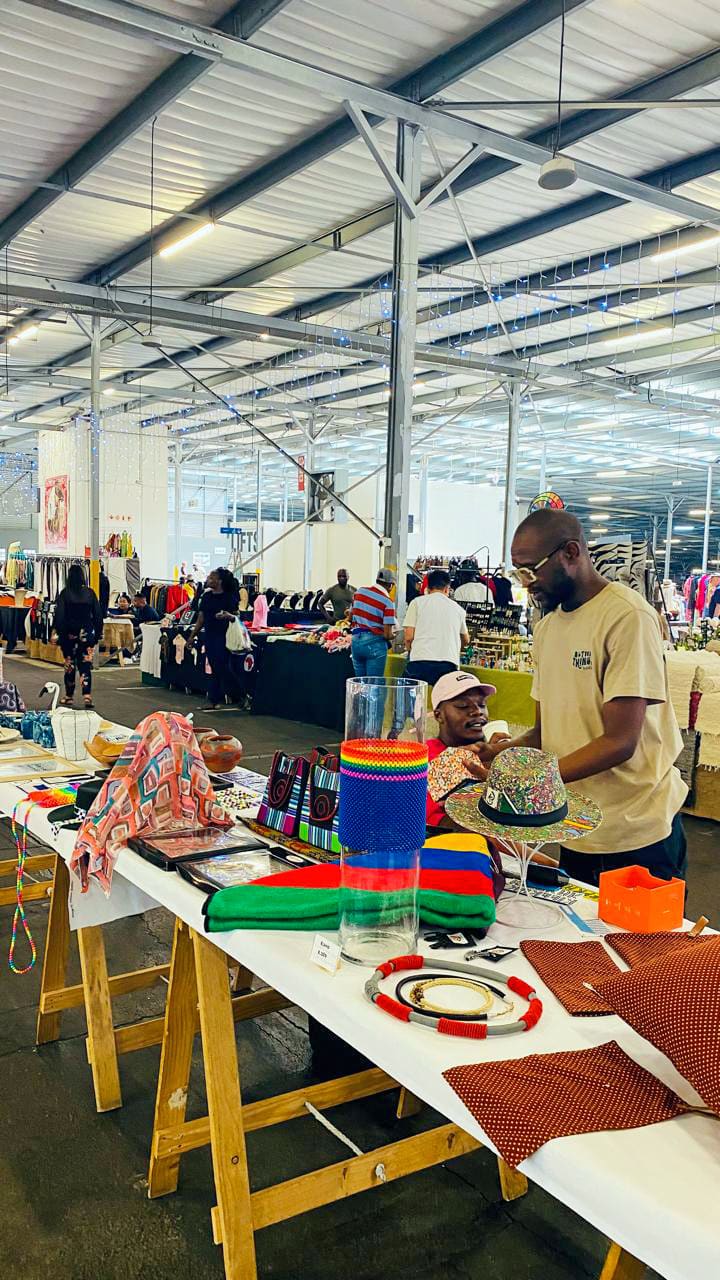 A man and woman talk at a vibrant stall with colourful handmade crafts and textiles, set inside a large indoor market. Other stalls with clothing and shoppers are visible in the spacious, industrial-style background.