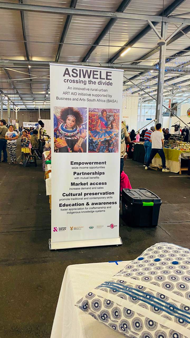 A large indoor market with people browsing stalls. In the foreground is a vertical banner for ASIWELE, a rural-urban art initiative, featuring two photos of women and text about empowerment, partnerships, and education. A patterned textile lies on a table nearby.