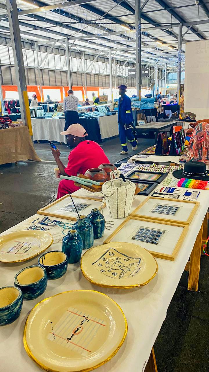 A craft market scene shows a table displaying handmade ceramics and framed art. Bowls and plates with blue patterns sit in front, with beaded jewellery and textiles further back. A person in pink sits behind the table; other shoppers browse stalls in a spacious indoor venue.