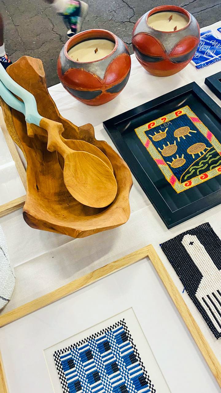 A display table features two round, red and beige candles, a carved wooden bowl holding two wooden utensils, and framed textiles with geometric and animal patterns in blue, yellow, black, and white. The surface is white and set indoors.