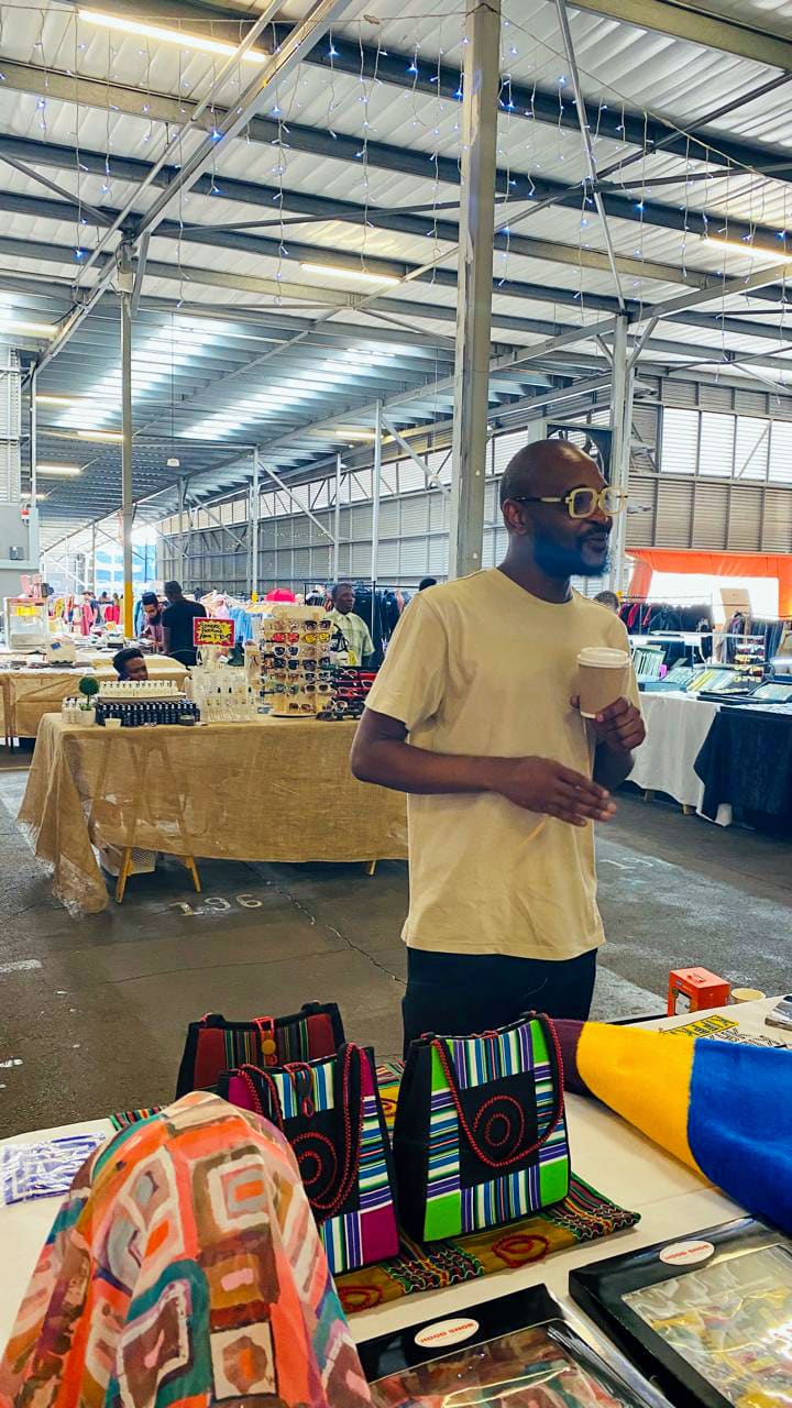 A man wearing sunglasses and a beige t-shirt holds a cup whilst standing behind a table with colourful patterned bags and fabrics at an indoor market, with stalls and fairy lights visible in the background.