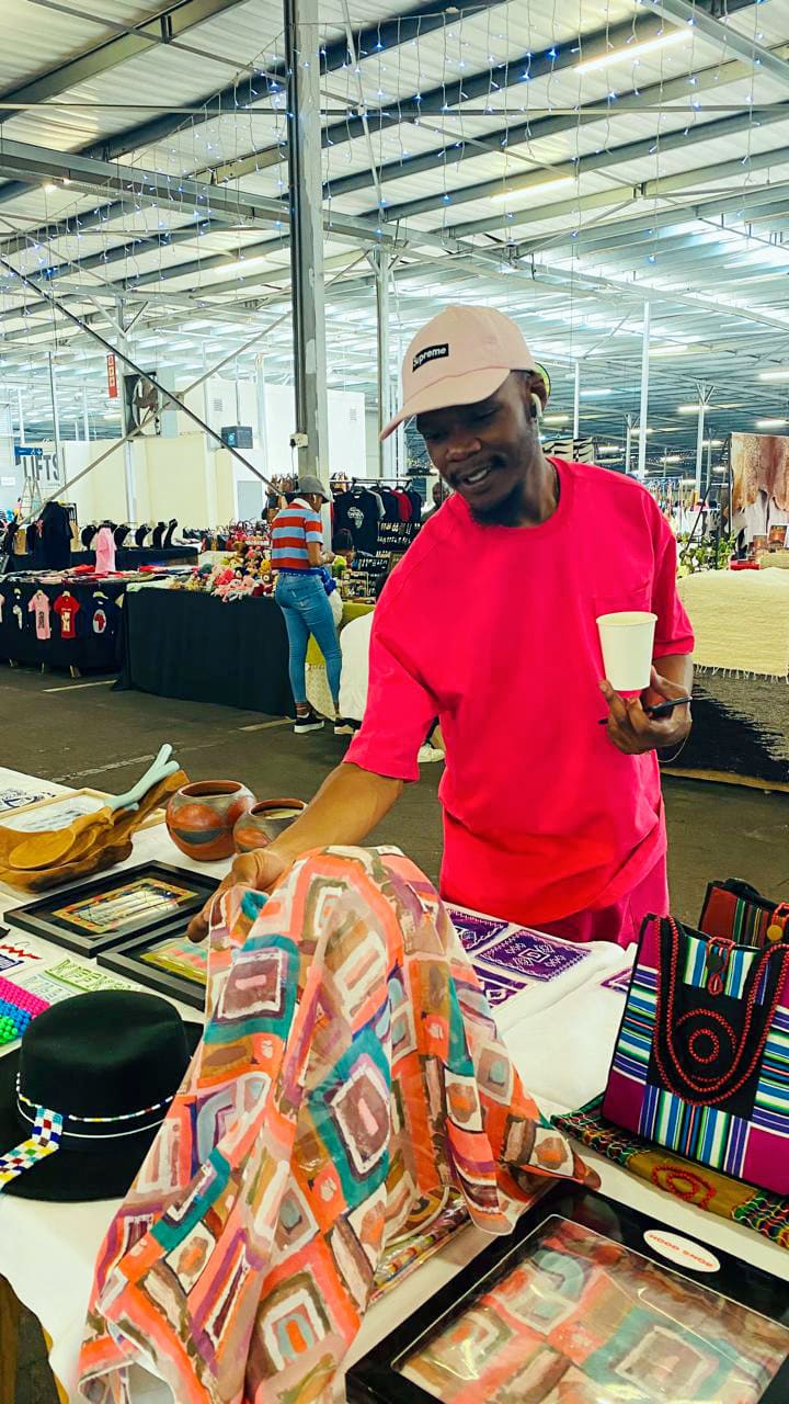 A man in a pink shirt and cap holds a white cup while browsing colourful handmade crafts at an indoor market stall. Artworks, textiles, and hats are displayed on the table. Other stalls and shoppers are visible in the background.
