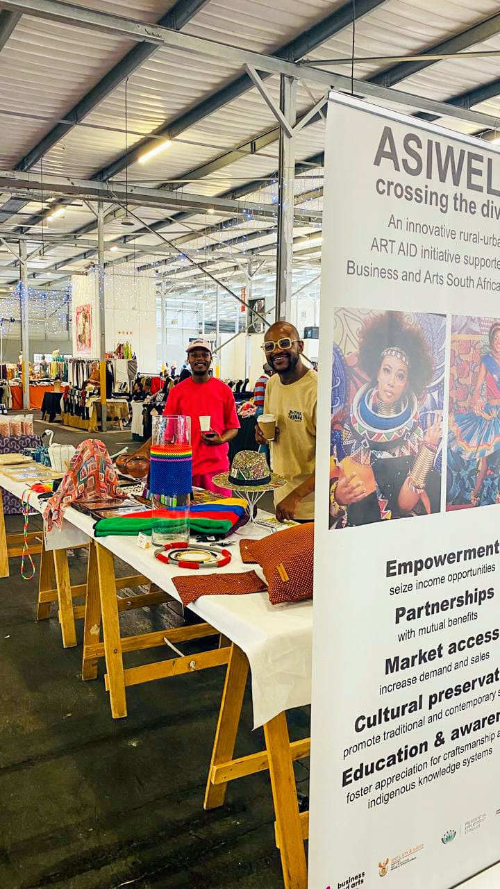 Two smiling people stand behind a colourful craft display at a market stall inside a large, bright building. The nearby banner reads “ASIWELE—crossing the divide” and highlights empowerment, partnerships, market access, and cultural preservation.