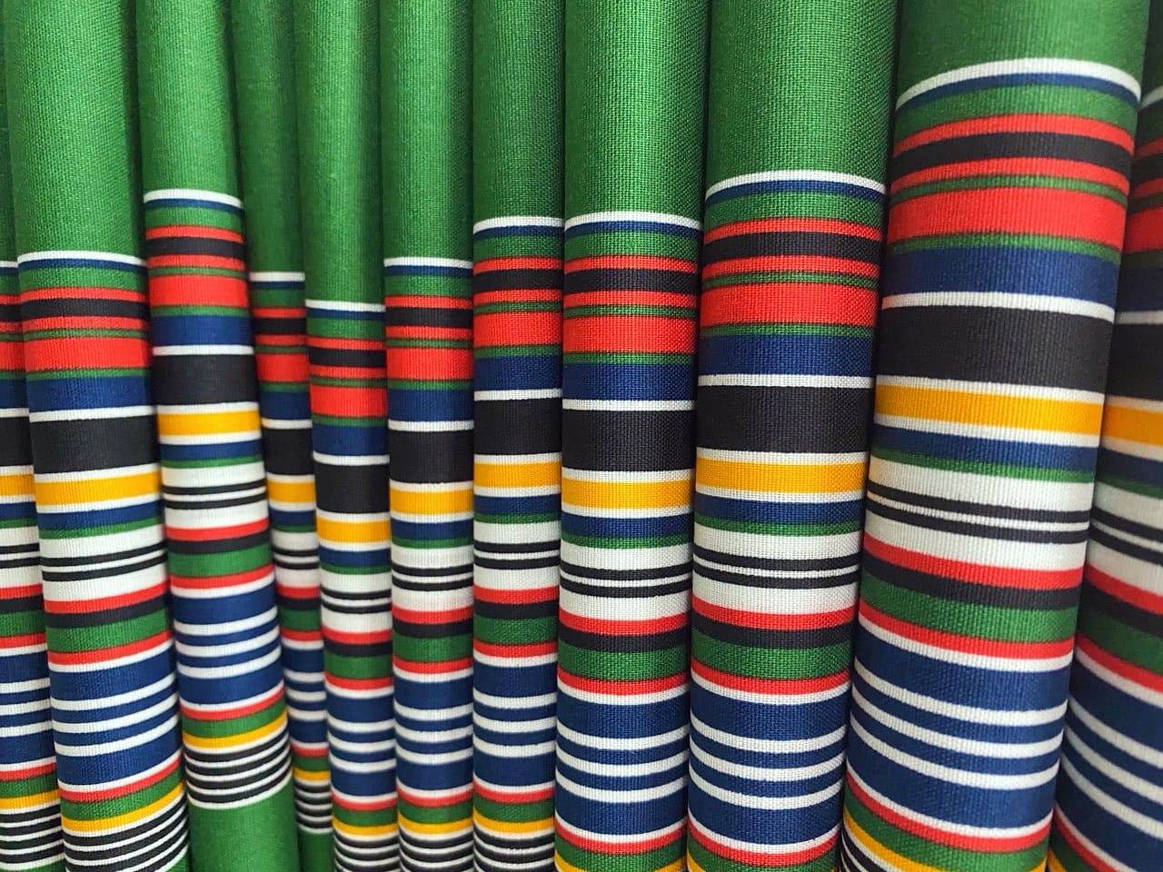 Rows of green fabric with horizontal stripes in red, blue, black, white, and yellow. The stripes vary in thickness and spacing, creating a colourful, repetitive pattern. The fabric appears to be vertically draped and closely arranged together.