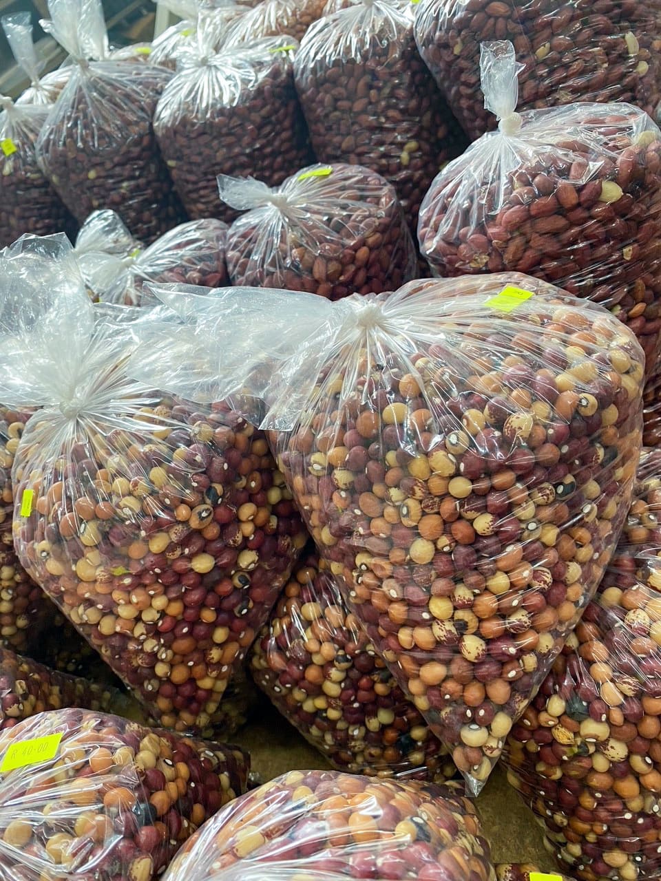 Large clear plastic bags filled with a mix of assorted dried beans and pulses are stacked in rows. The beans inside are of various colours including brown, beige, and red. Each bag has a small yellow price sticker attached. The bags are tightly packed together.