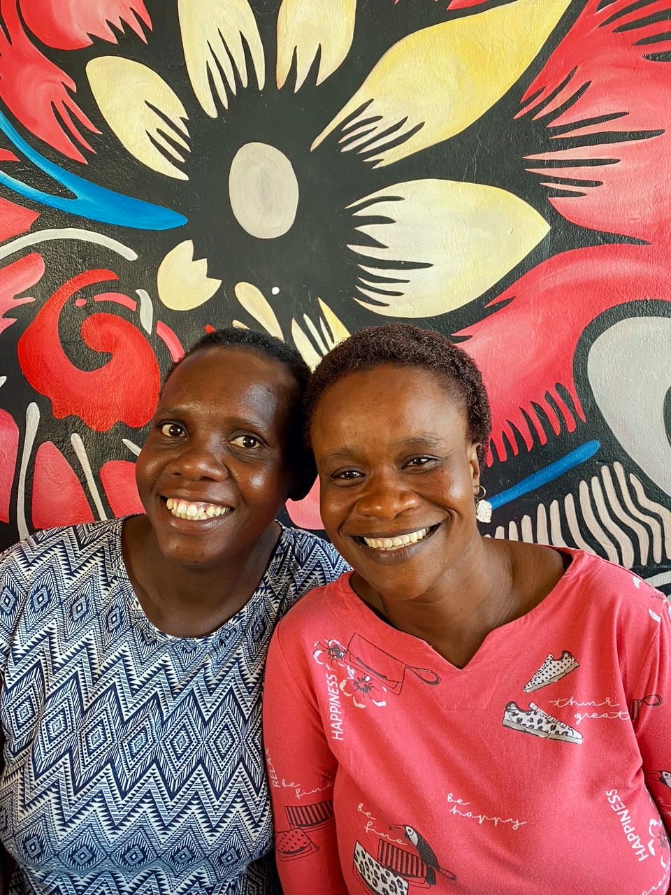 Two women sit close together smiling, in front of a bright, colourful mural featuring a large, abstract flower design in yellow, red, white, and black. One wears a blue patterned shirt; the other wears a pink shirt with white writing and illustrations.