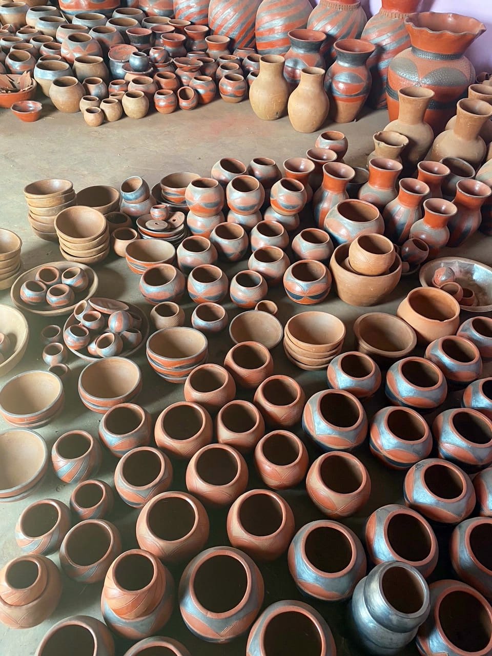 Rows of handmade clay pots, bowls, and vases in various sizes and shapes are arranged on the ground. Most are reddish-brown, some with blue and grey painted patterns. The pottery is grouped by type, filling the image, and the background shows more stacked vessels.
