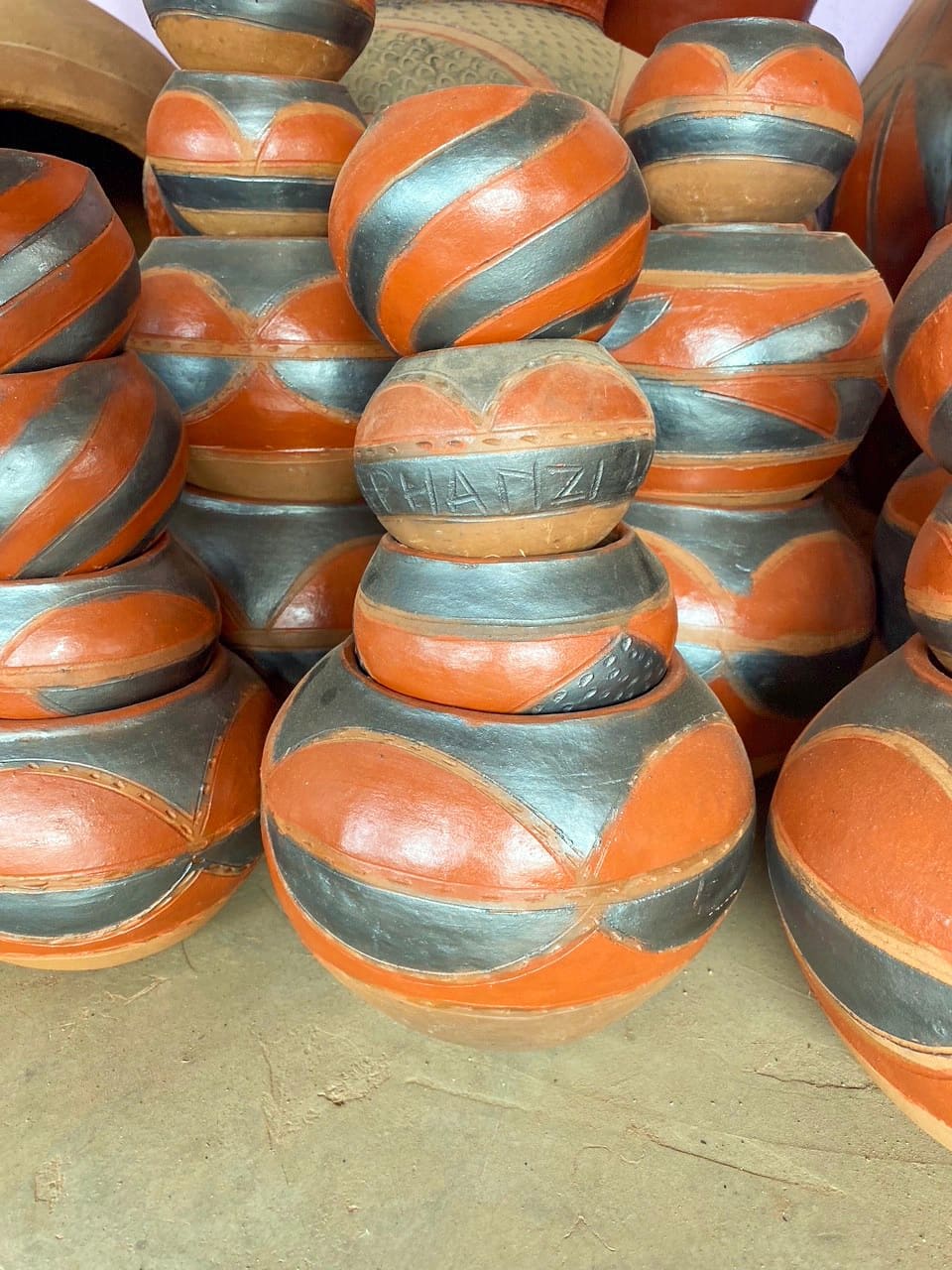 A group of stacked, round clay pots with smooth surfaces, decorated in curved orange-brown and black patterns. The pots are arranged closely together on a brown surface, with etched markings visible on one of the upper pots.