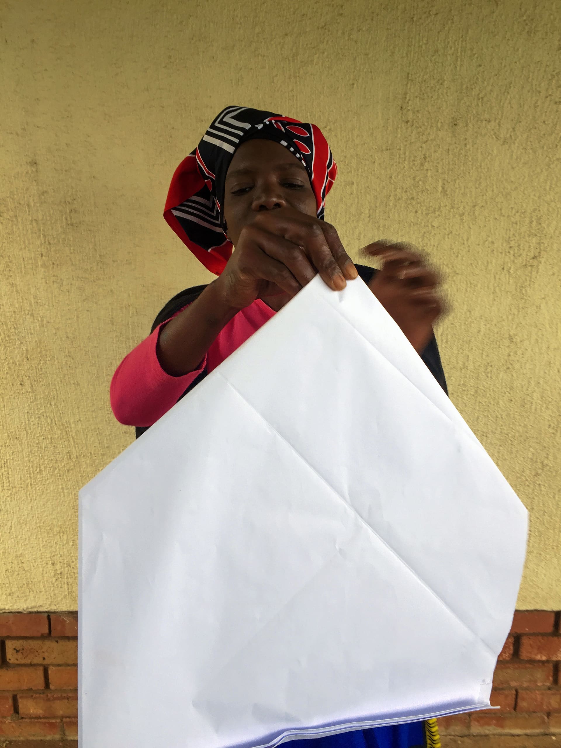 A woman wearing a colourful headscarf and pink sleeves folds a large white sheet of paper. She stands in front of a yellow wall with a brick border at the bottom, looking focused on the paper in her hands.