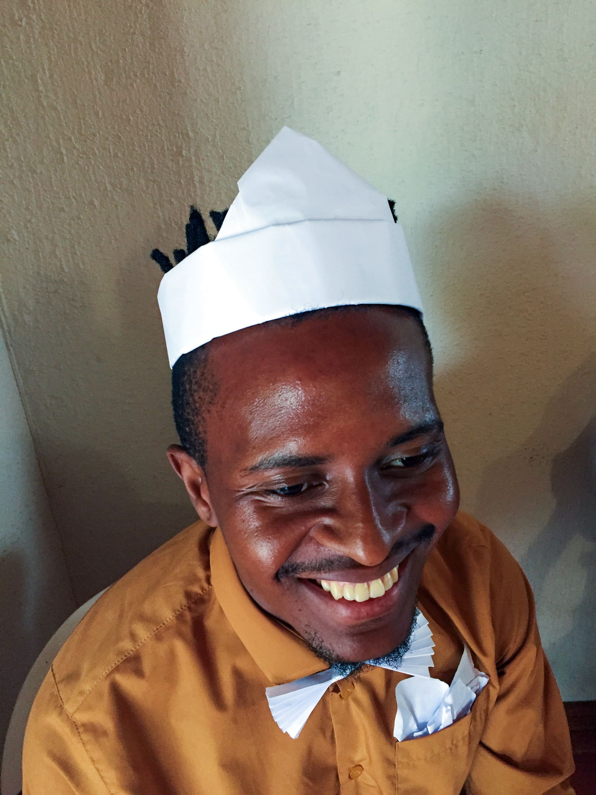 A smiling man wearing a mustard-coloured shirt, white paper bow tie, and a white paper hat sits indoors. His shirt pocket also has a folded white paper. The wall behind him is cream-coloured, and warm light highlights his face and joyful expression.