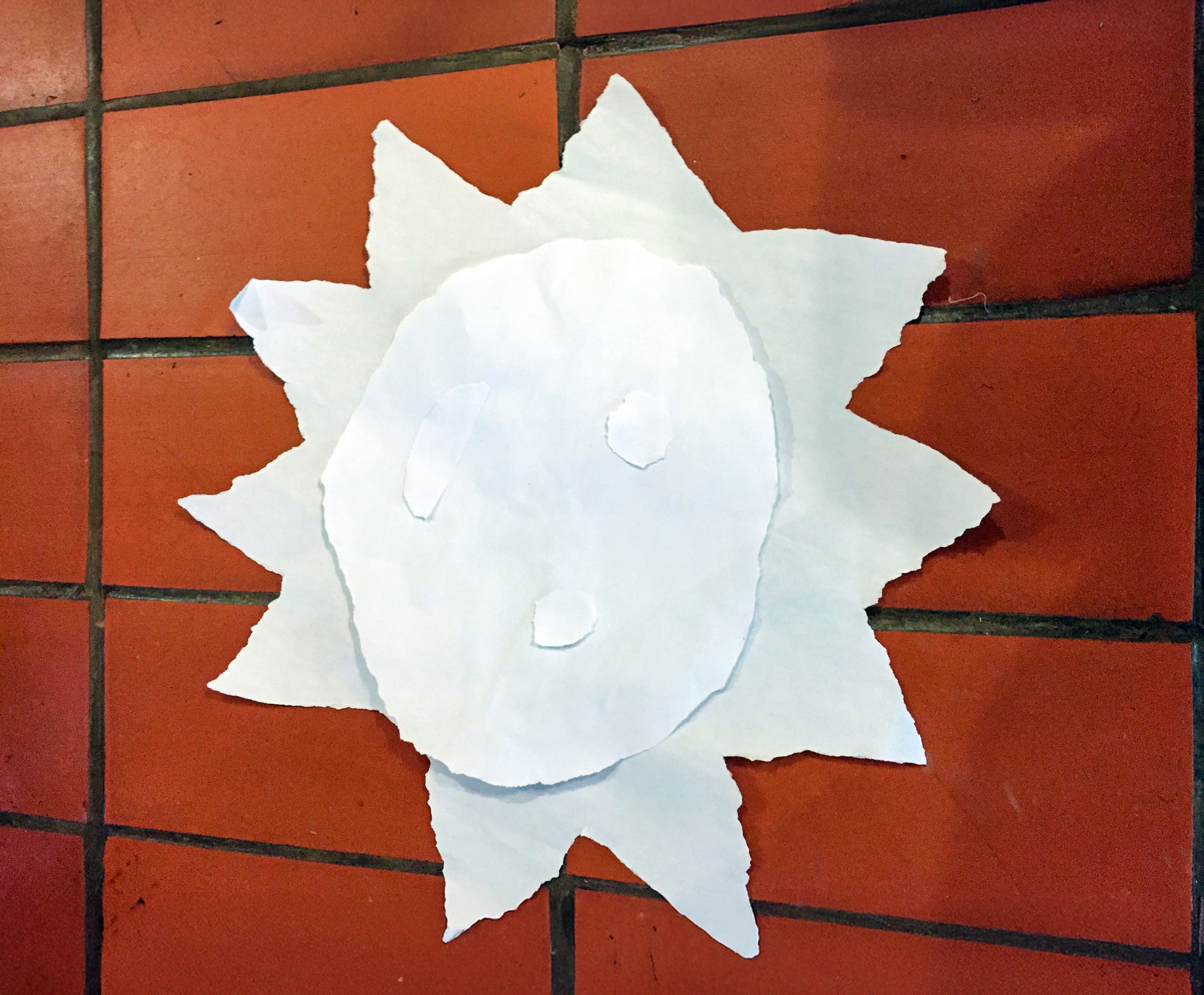 A sun shape made from torn white paper with a round centre and jagged rays is attached to a wall of reddish-orange square tiles. The sun has two small ovals for eyes and a round mouth, also created from torn white paper.