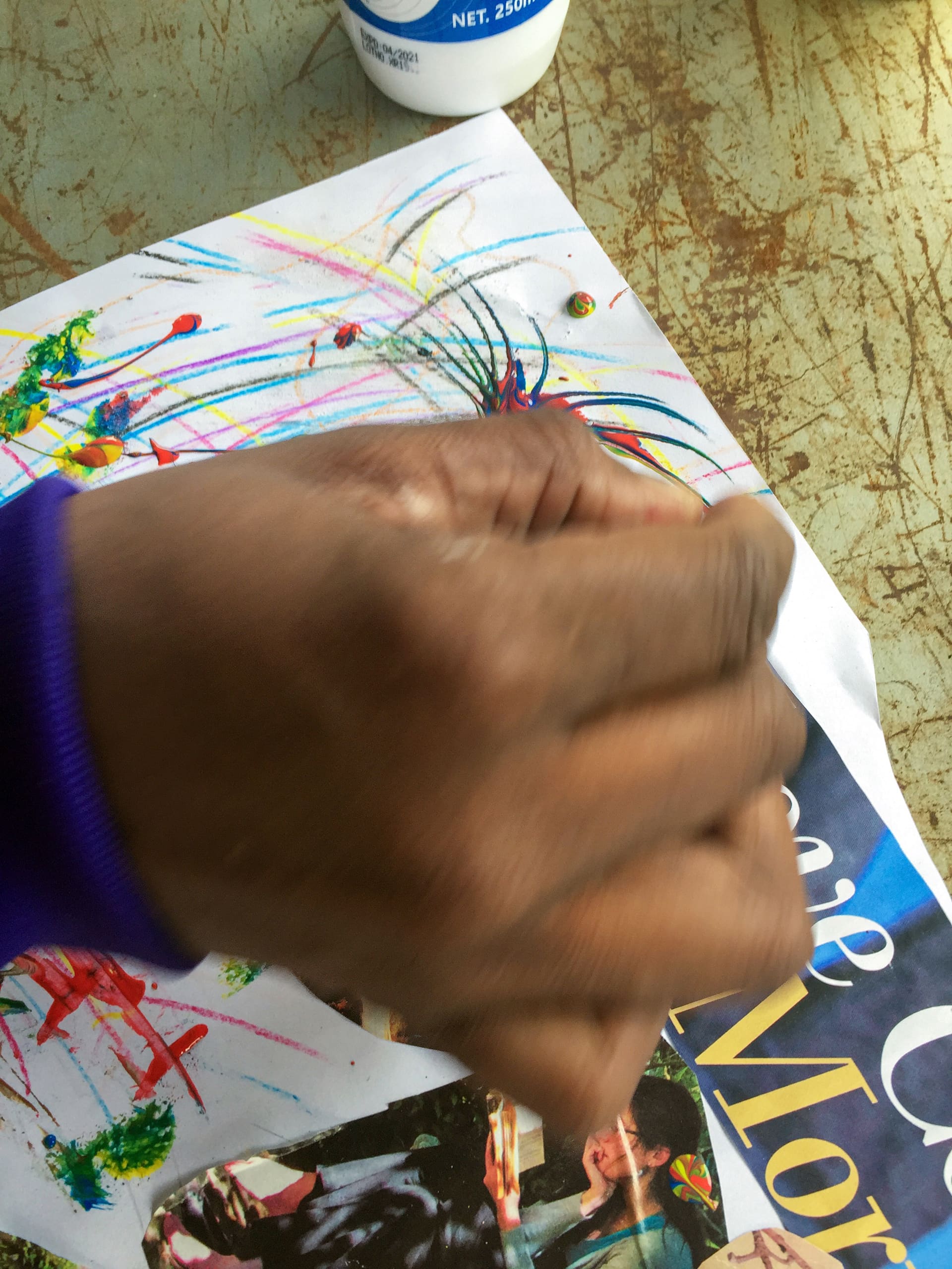 A hand in motion blurs over a piece of white paper with colourful crayon scribbles and magazine cut-outs. Part of a blue sleeve is visible. At the top, there’s a partially seen white glue bottle on a worn, greenish surface.
