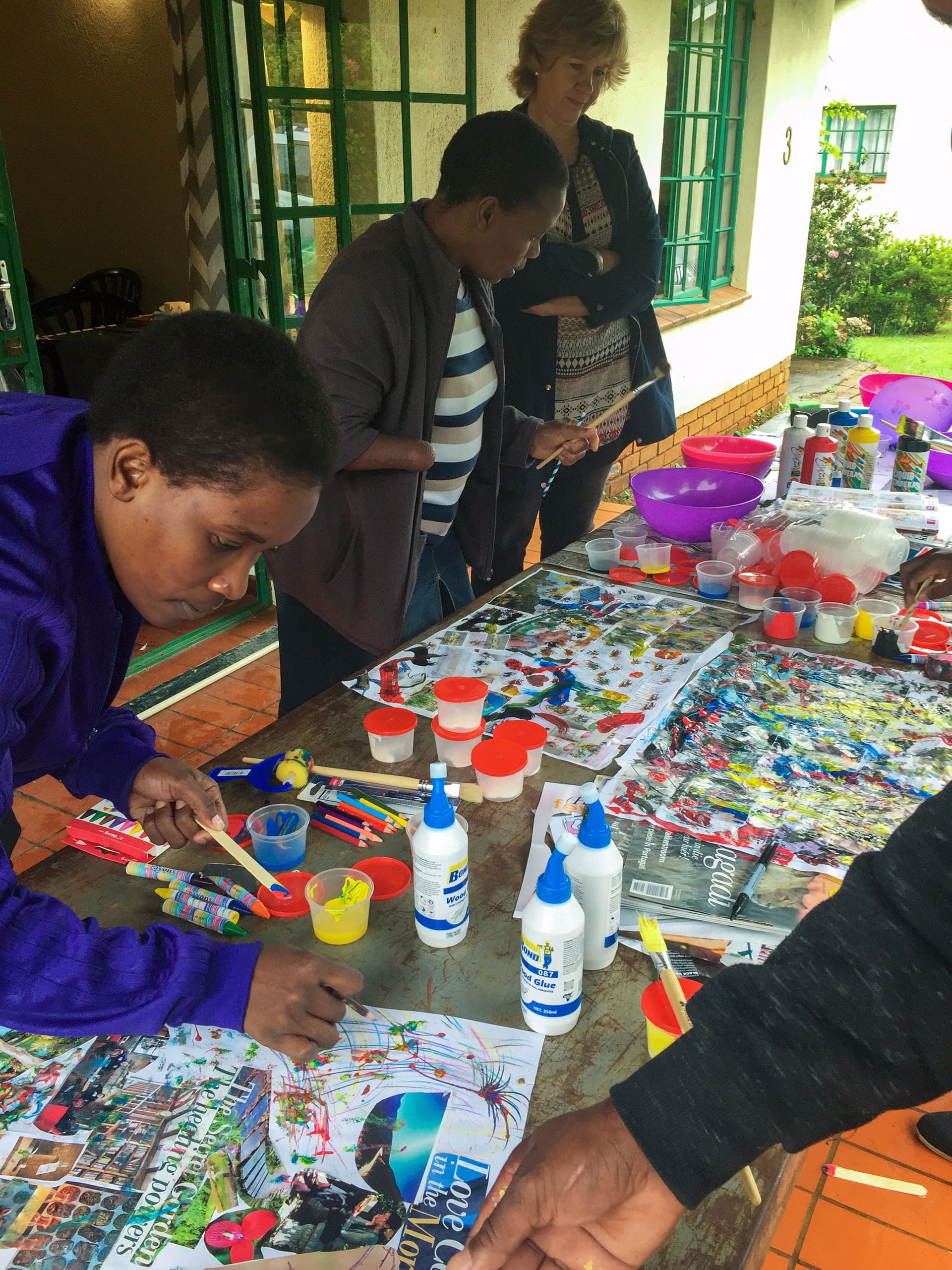 Three people work on colourful arts and crafts at a table covered with magazines, glue, paint, and supplies. Two are focused on their projects, while a third person stands watching. The setting appears to be outdoors, next to a building with green window frames.
