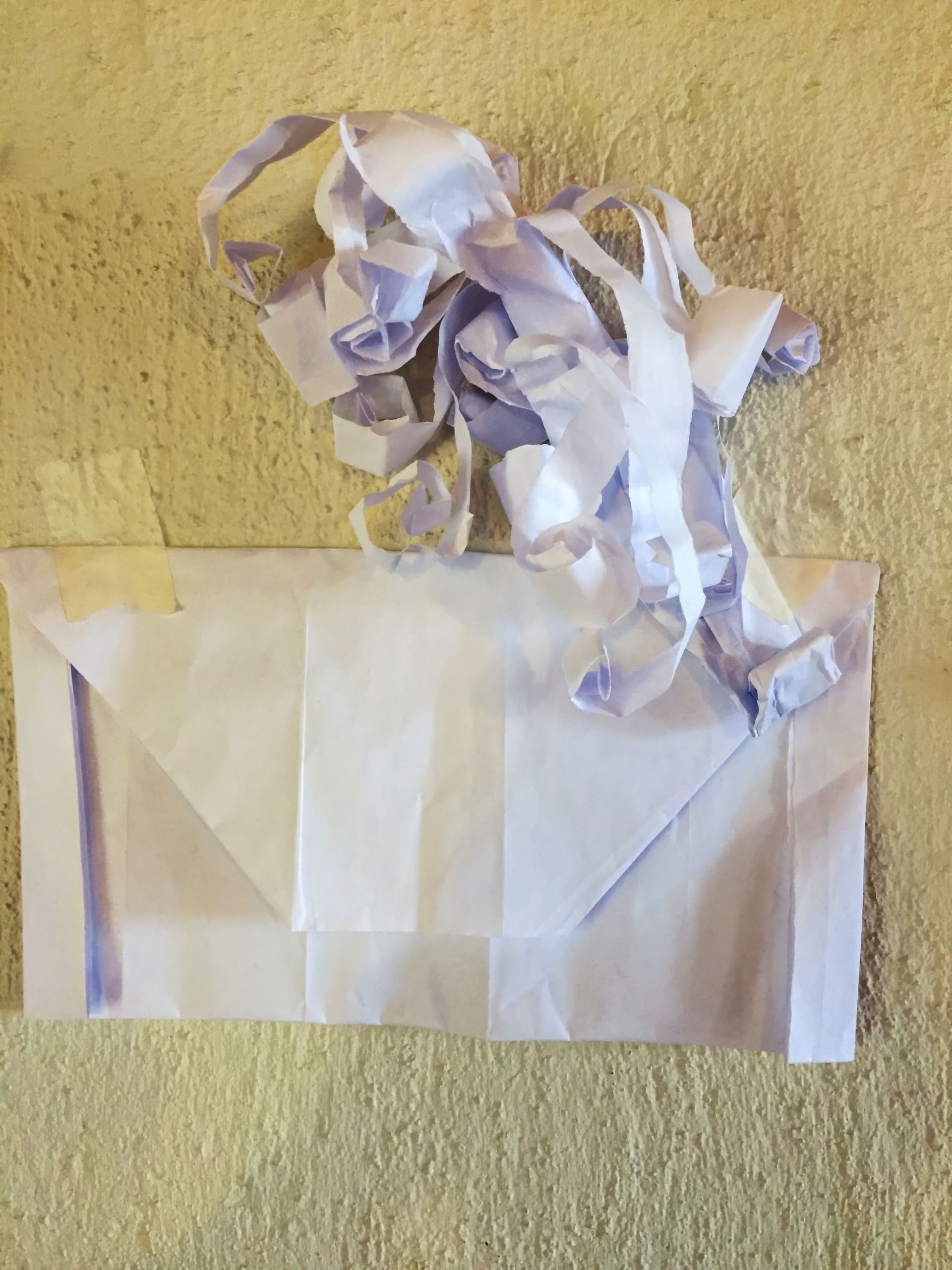 A handmade white paper handbag is taped to a textured beige wall. The handbag has a flap and two handles made from curled paper strips, appearing crumpled and uneven. The edges and corners are held together with visible pieces of tape.
