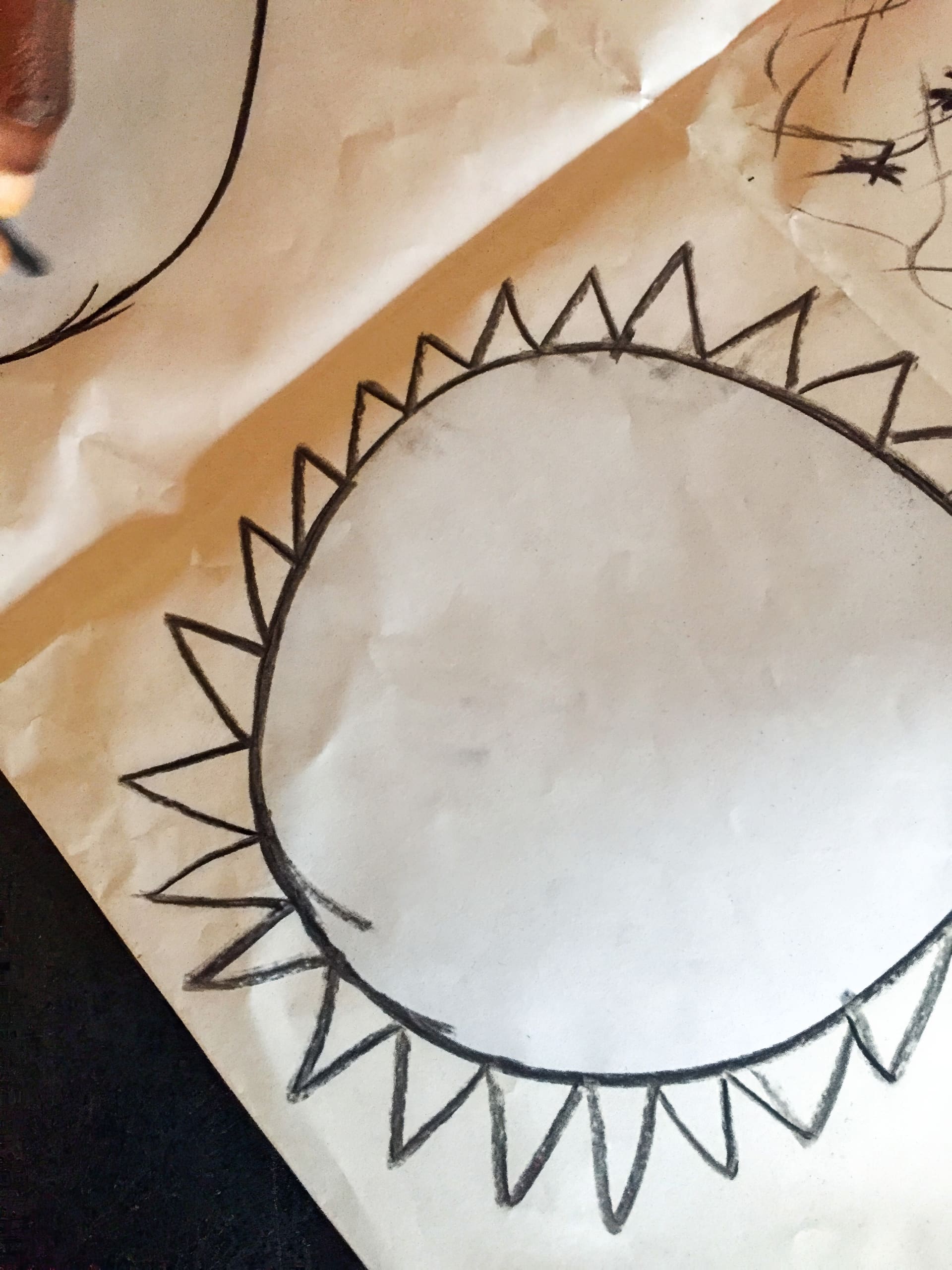A piece of paper shows a large circle drawn in black crayon with many triangle shapes around the edge, resembling a sun. There are extra scribbles and a partial curved shape visible on the top left, and a hand holding a crayon at the top left corner.