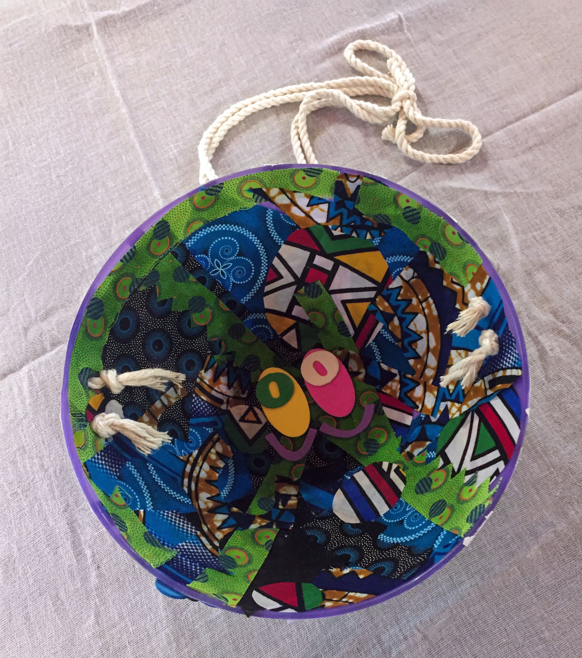 A colourful circular handbag with a braided rope handle, featuring a vibrant patchwork of blue, green, black, and purple patterns, and two overlapping button-like decorations on top. The bag rests on a light beige fabric surface.