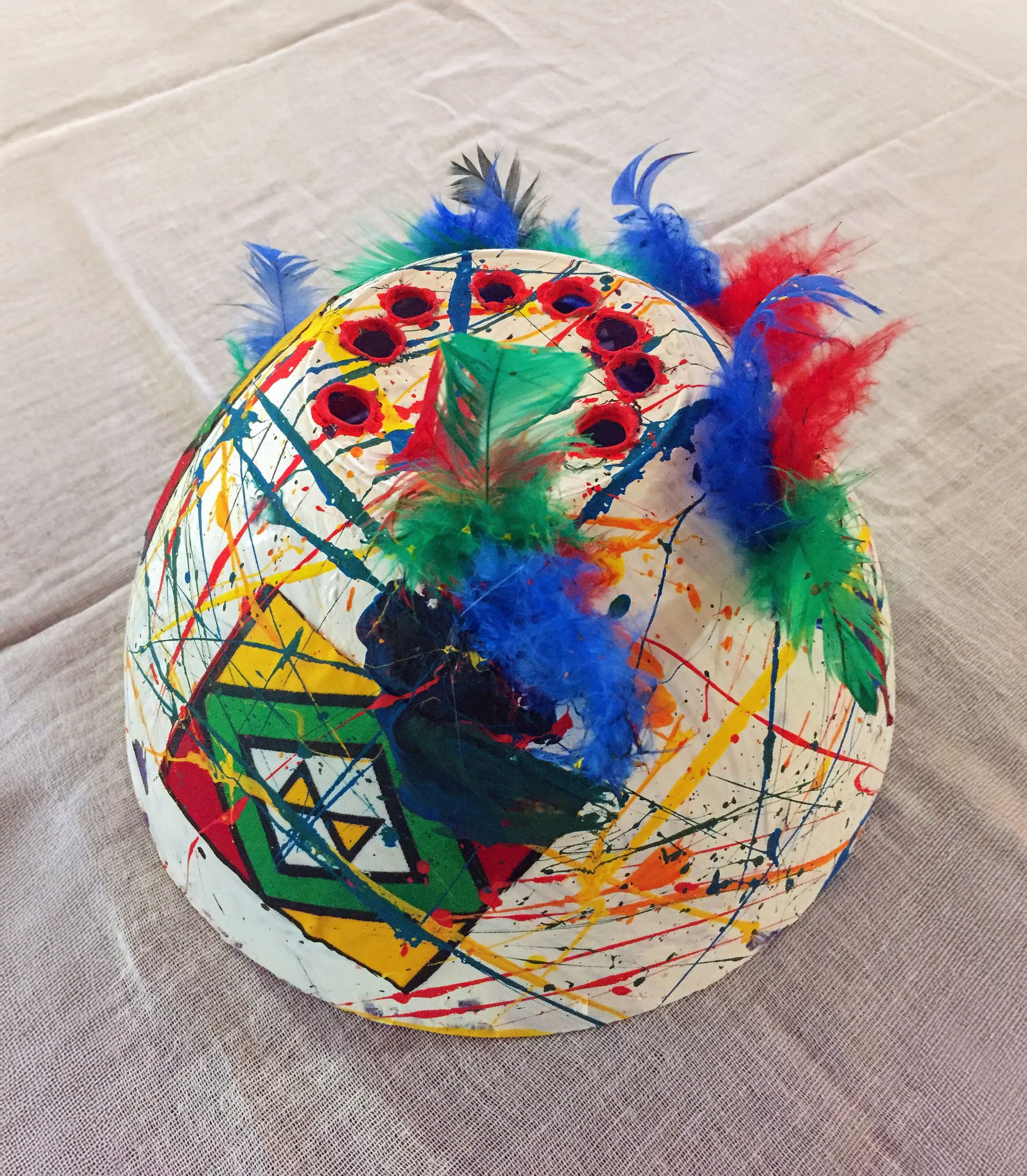 A white papier-mâché mask decorated with colourful paint splatters, geometric shapes, and circular red gems. Blue, green, and red feathers are attached on top, giving the mask a festive and artistic appearance. The mask sits on a textured cloth surface.