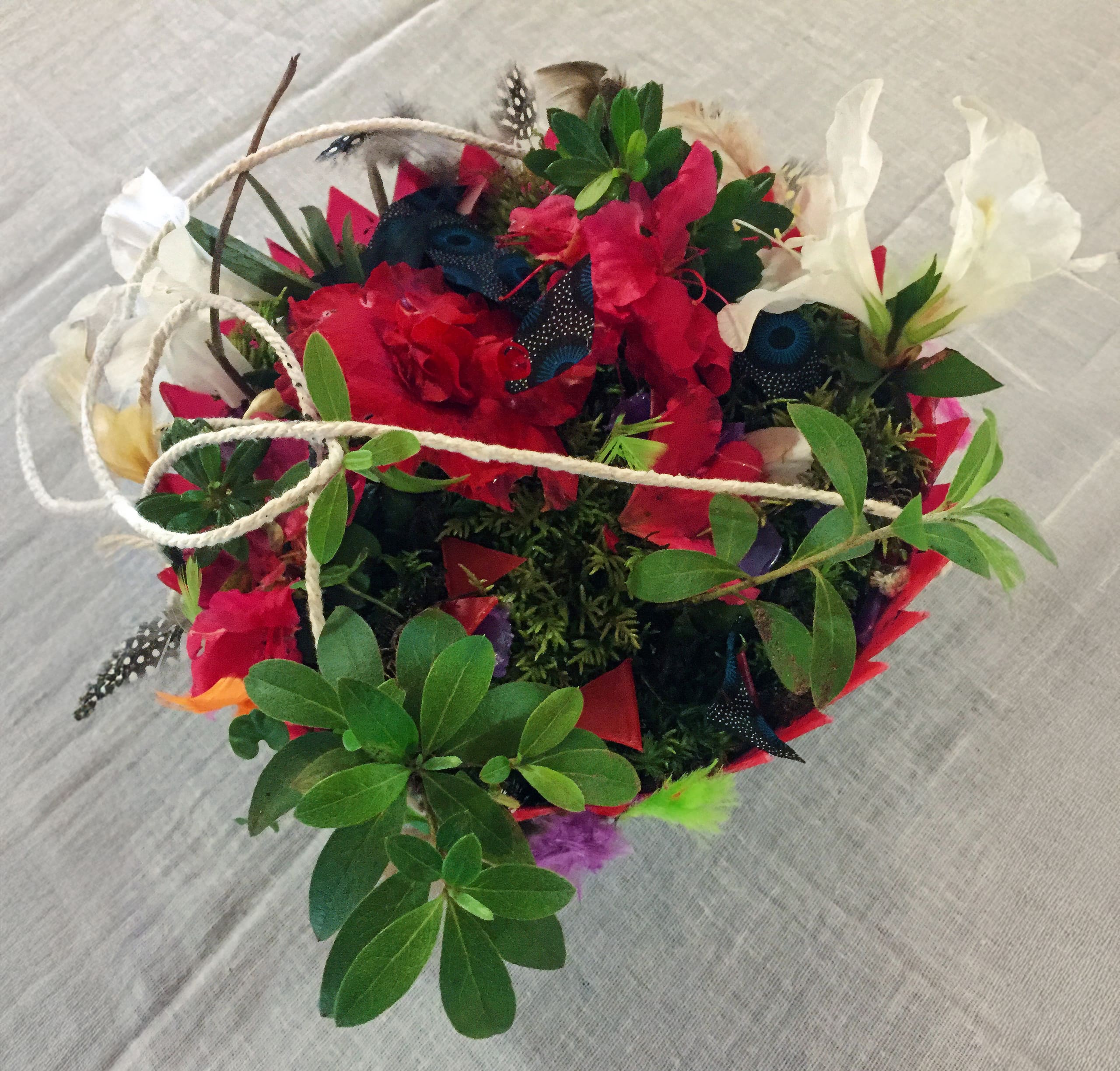 A vibrant floral arrangement in a round container, featuring red, white, and pink flowers, green leaves, feather accents, and a white string arranged on top. The basket sits on a light grey textured fabric background.