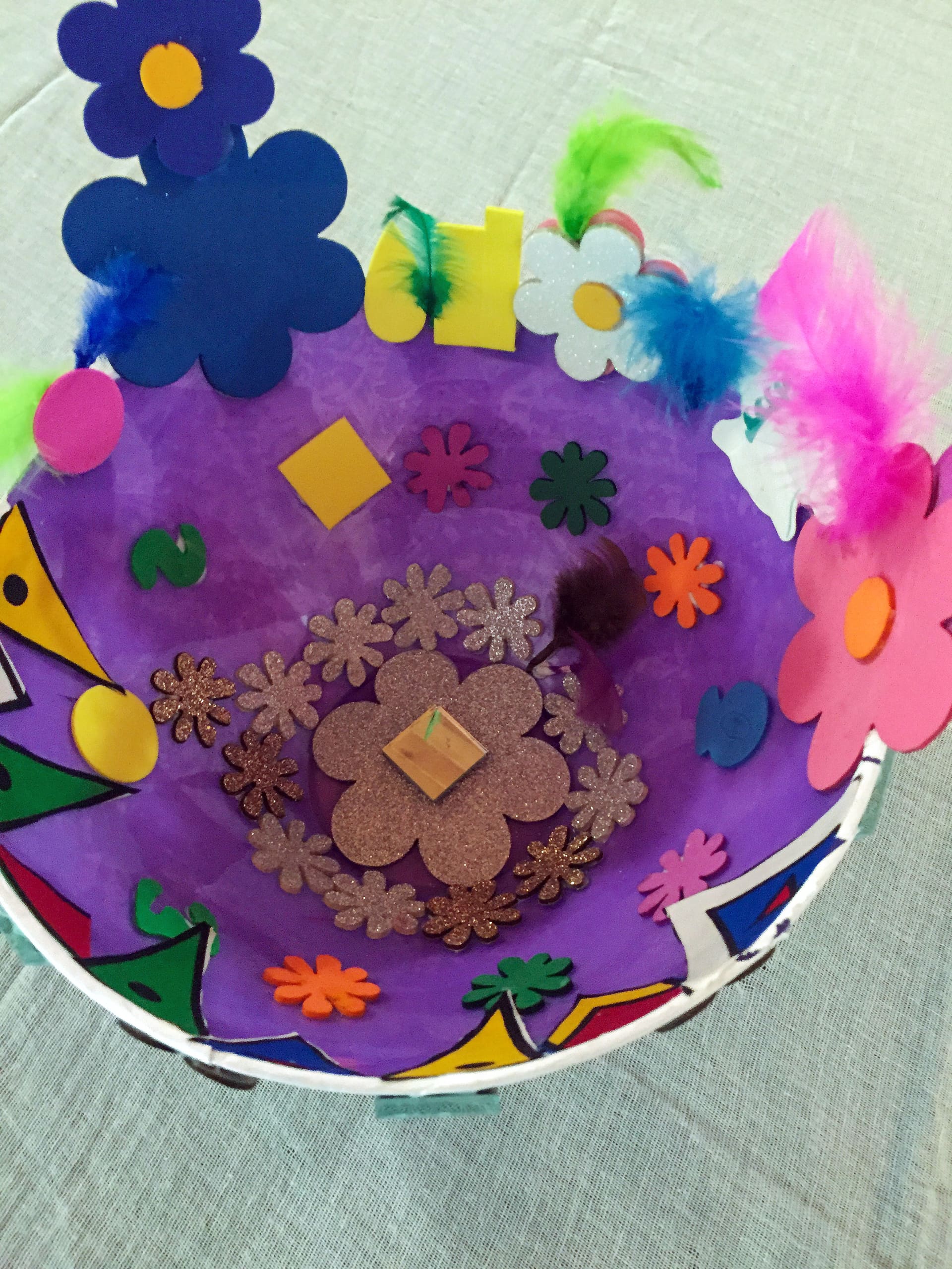 A colourful, handmade bowl decorated with large foam flowers, glittery flower shapes, feathers, and small foam shapes. The bowl’s interior is mostly purple, with an array of bright decorations inside and around the rim, set on a pale textured surface.