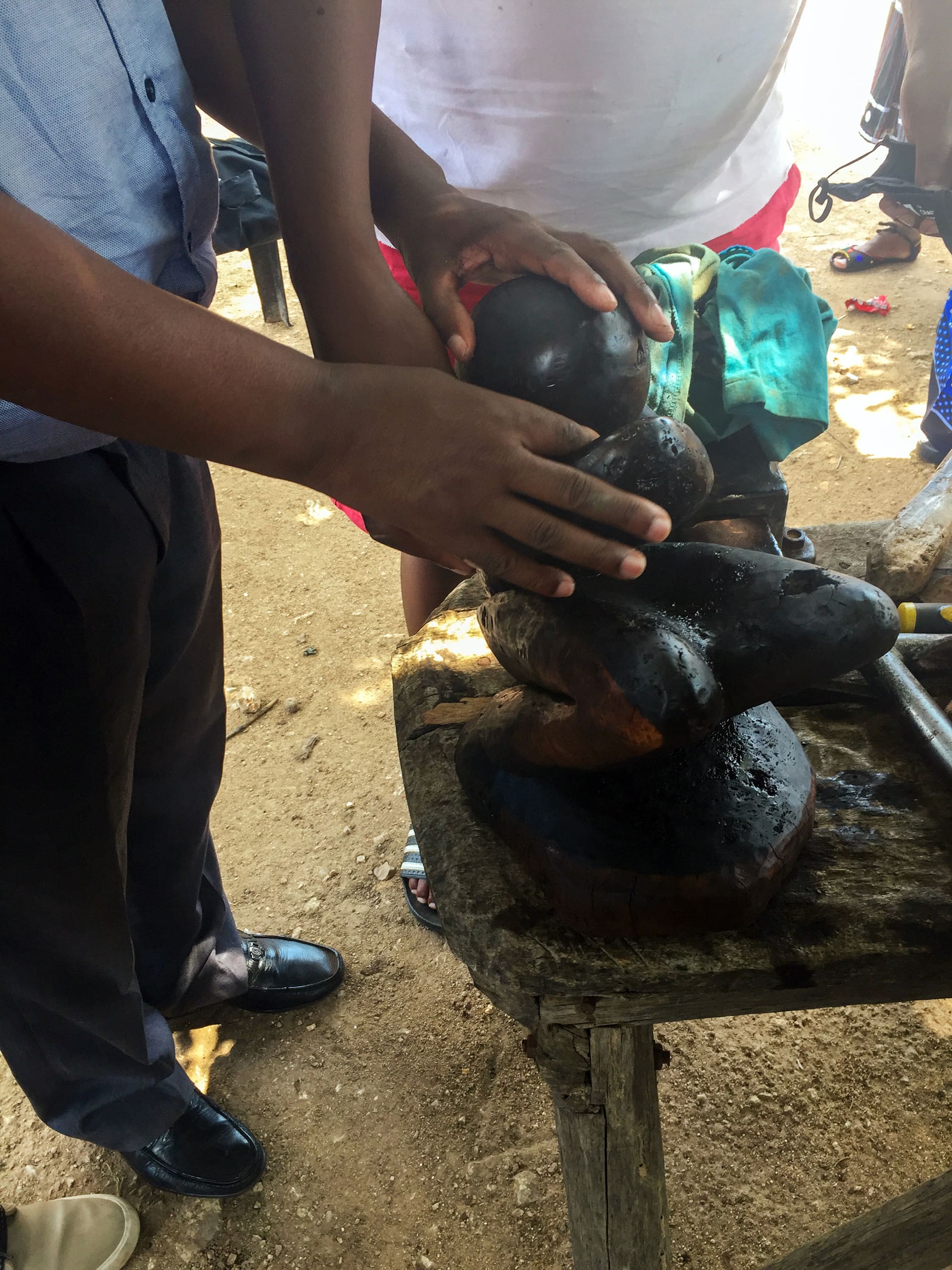 Three people’s hands are touching a dark, carved wooden sculpture on a rustic wooden table outdoors. One person wears formal trousers and shiny shoes, another is barefoot. The scene suggests a collaborative or instructive activity, possibly related to art or craftsmanship.