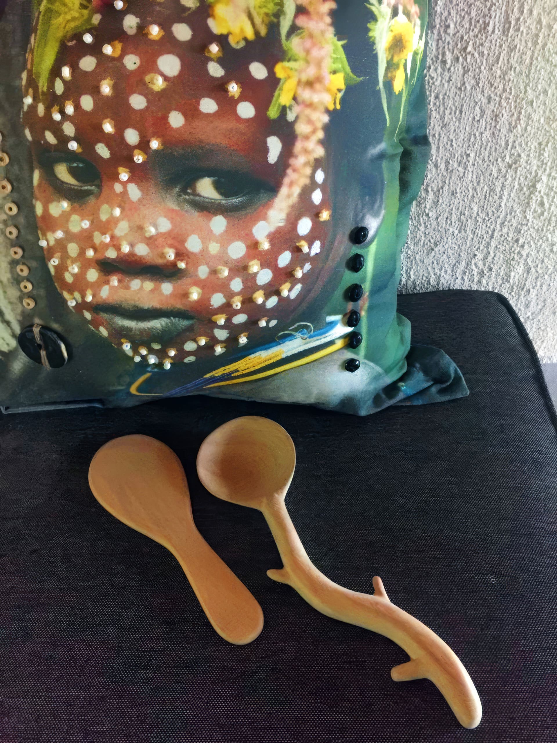 A decorative cushion with an image of a child’s face painted with white dots and adorned with yellow flowers rests against a textured wall. In front, two wooden spoons—one simple and one shaped like a tree branch—lie on a dark fabric surface.