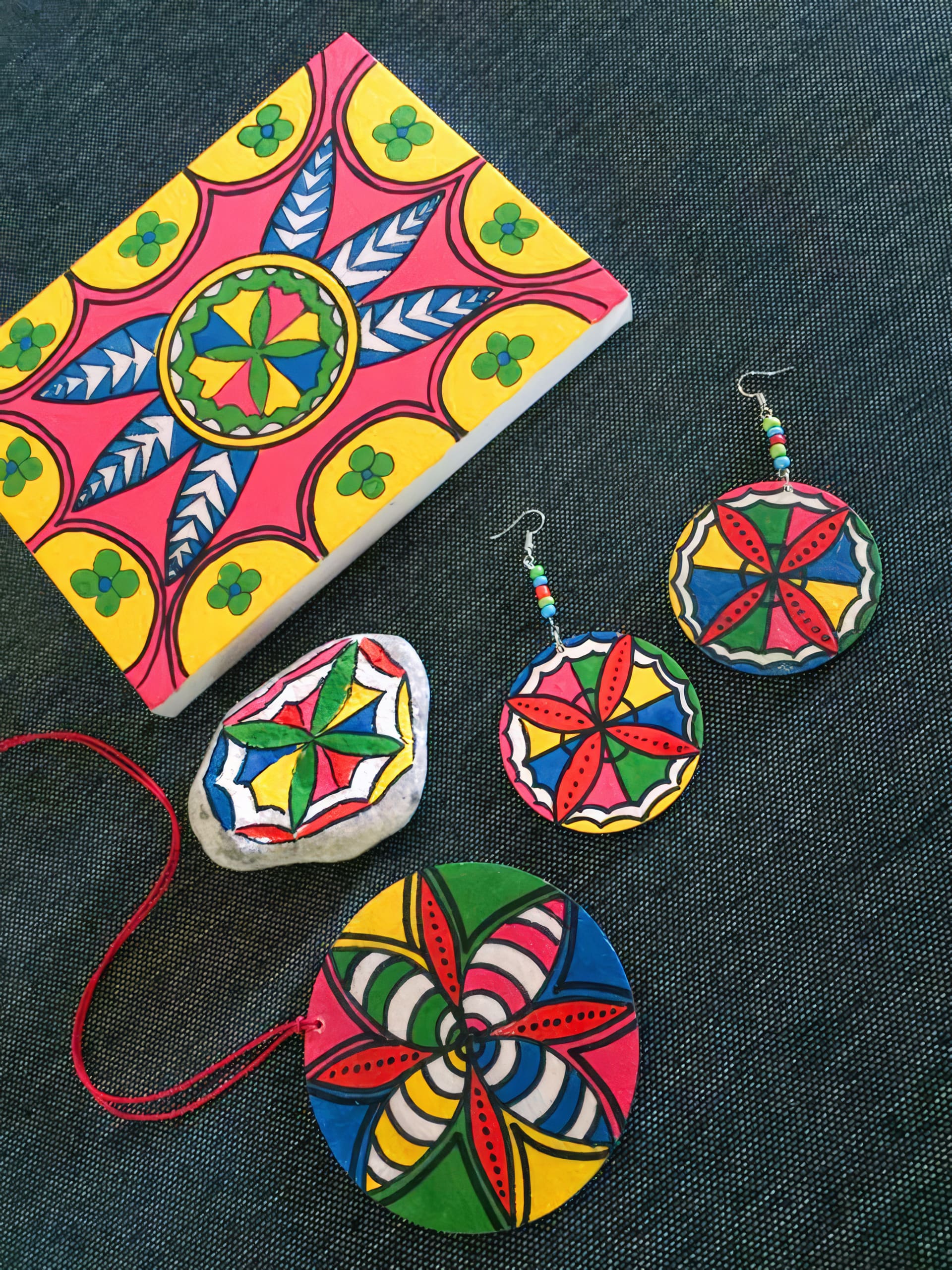 A colourful notebook, two round earrings, a pendant, and a painted stone rest on dark fabric. All feature bold, geometric folk designs in red, yellow, green, and blue, with repeating star and petal shapes outlined in black.