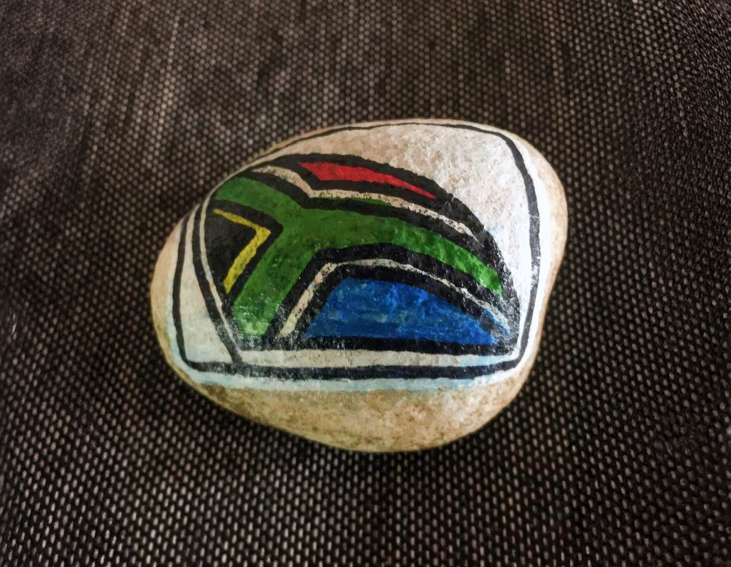 A smooth, oval stone features a hand-painted design resembling the South African flag, with sections of green, black, yellow, red, blue, and white outlined in black. The stone rests on a textured, dark fabric background.