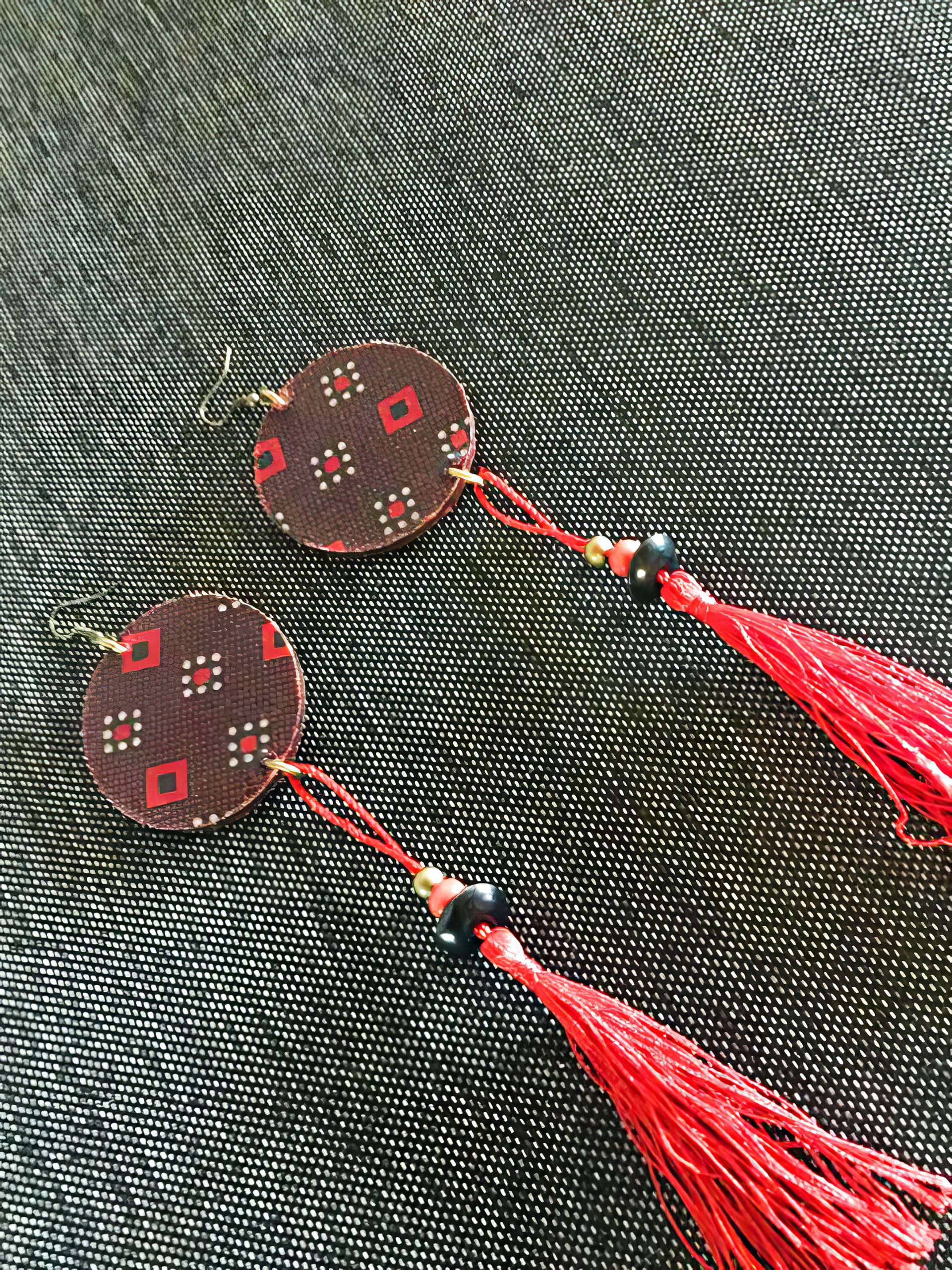 A pair of handmade earrings with round, patterned discs featuring red and white diamond shapes, black beads, gold accents, and long, bright red tassels, displayed on a textured dark grey fabric background.