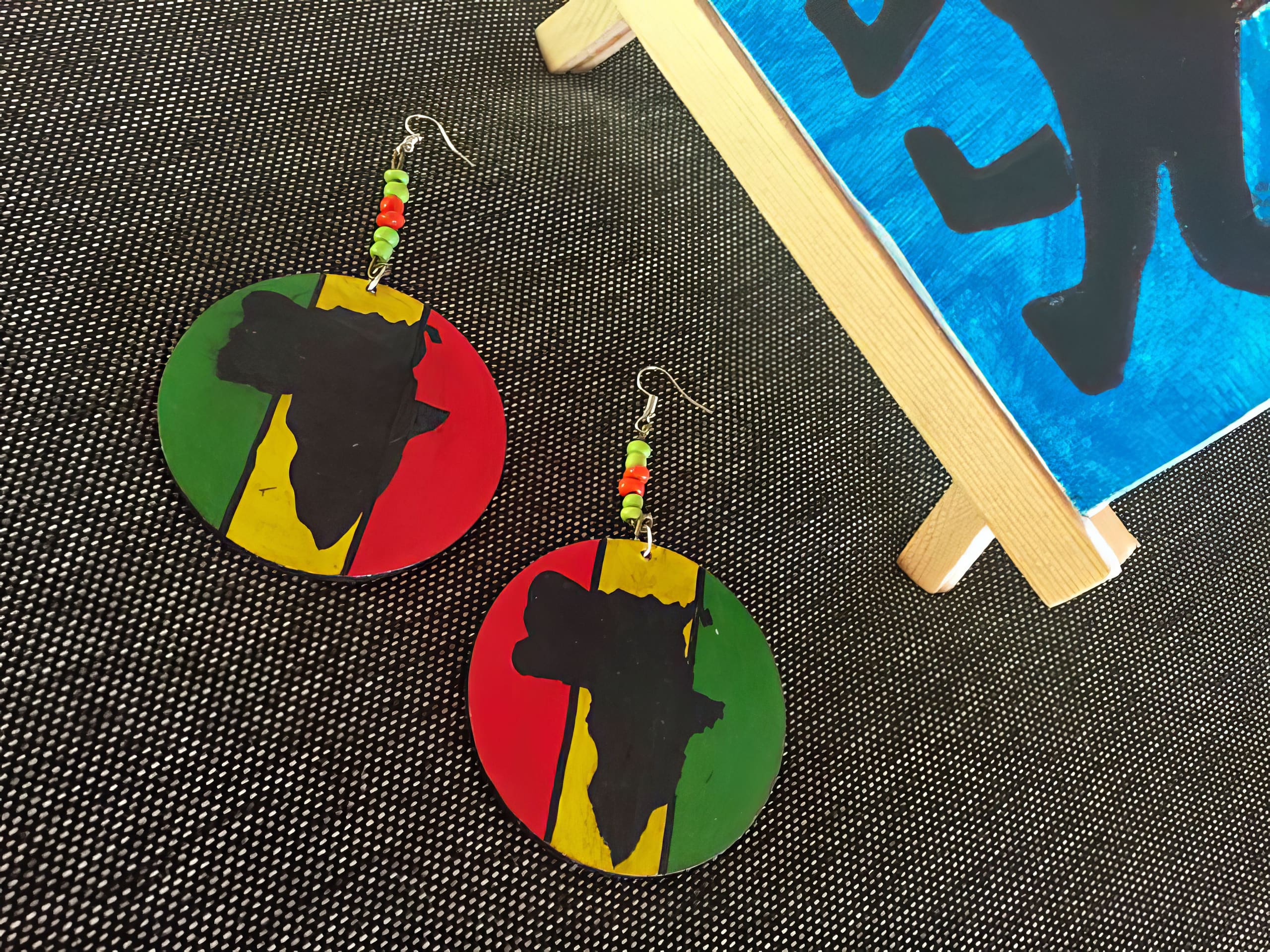 Two round earrings with the African continent silhouette in black over green, yellow, and red stripes. Each earring has a beaded hook in matching colours. Part of a blue canvas with a black design and wooden edge appears on the right. Background is dark fabric.