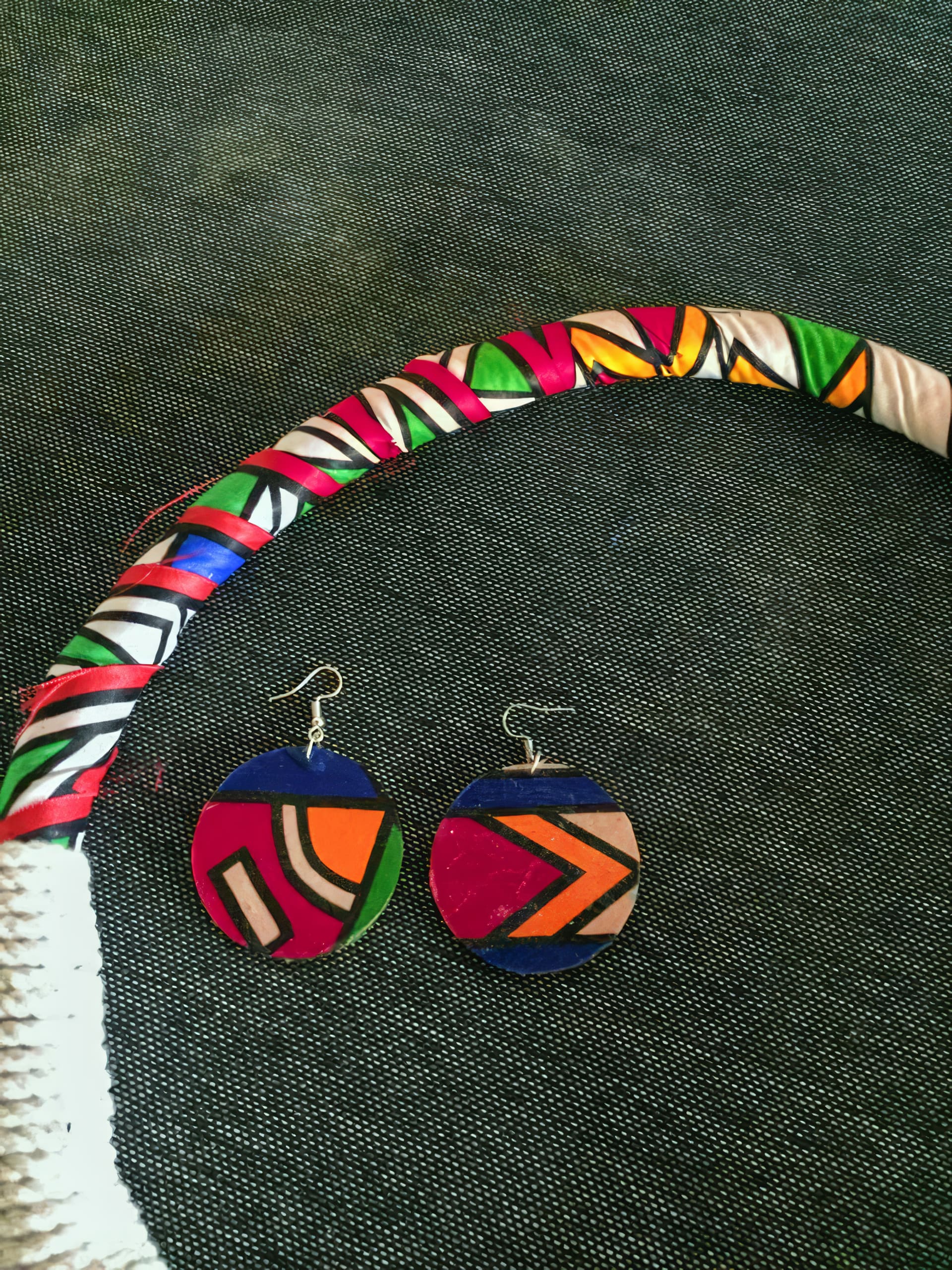 A colourful hoop wrapped with patterned fabric lies on dark textured fabric. Below it are oval earrings with bold, geometric designs in blue, red, orange, pink, and green, echoing the vibrant hoop colours.