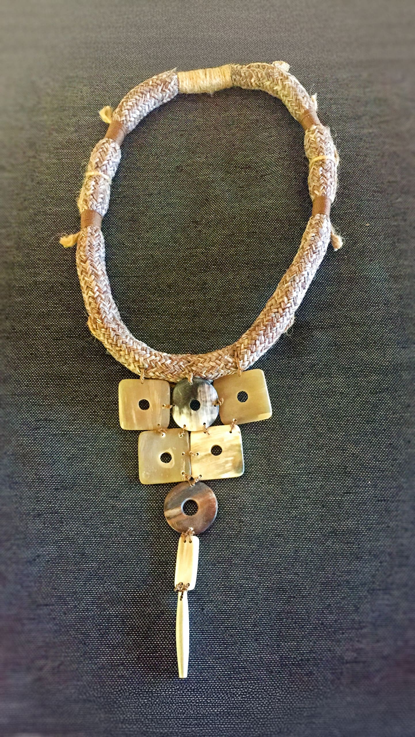A chunky, textured necklace with a thick, braided tan cord features a pendant made of rectangular and circular pieces in varying shades of cream and brown, ending in a long, pointed charm. The necklace is displayed on a dark textured background.