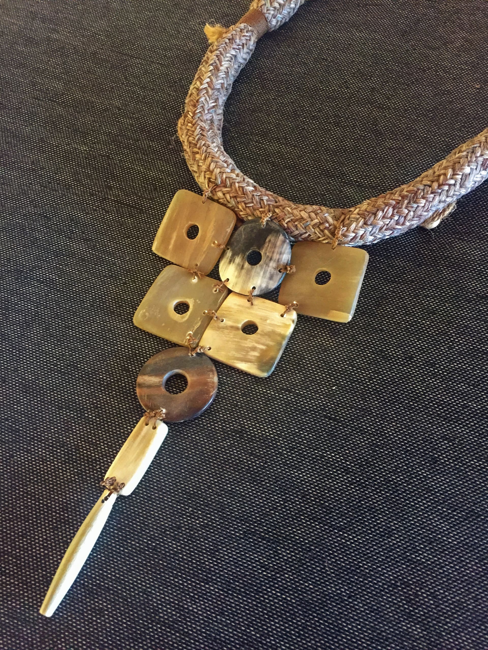 A necklace with a plaited, light brown cord features five square and circular horn or shell pieces, each with a central hole, arranged in a V shape. Below, a long, tapered pendant extends downward. The background is dark fabric.
