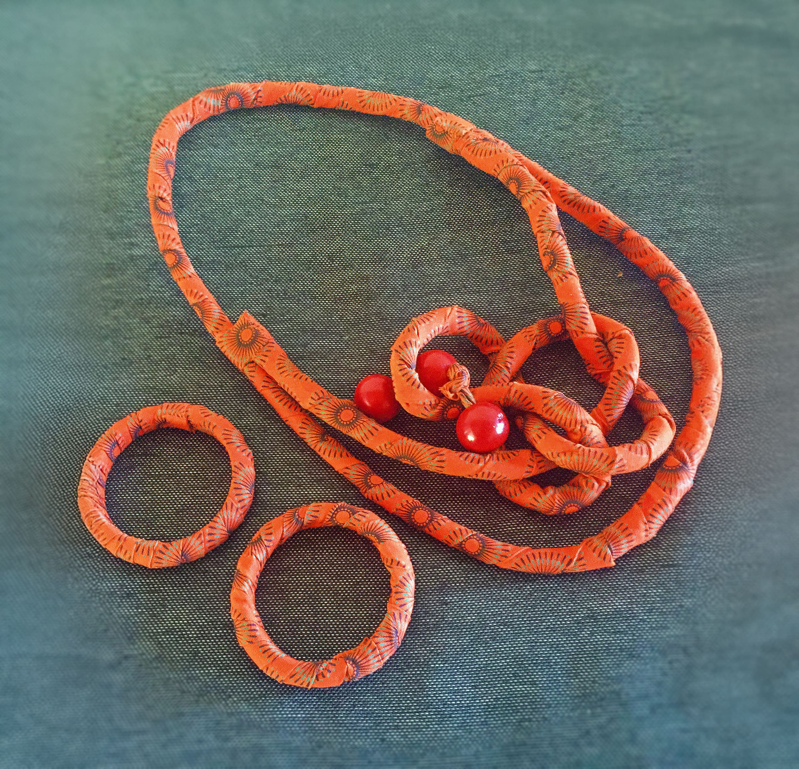A set of handmade jewellery on a textured teal surface: an orange patterned fabric necklace with loops and three red beads, and two matching orange fabric bangles placed beside it.