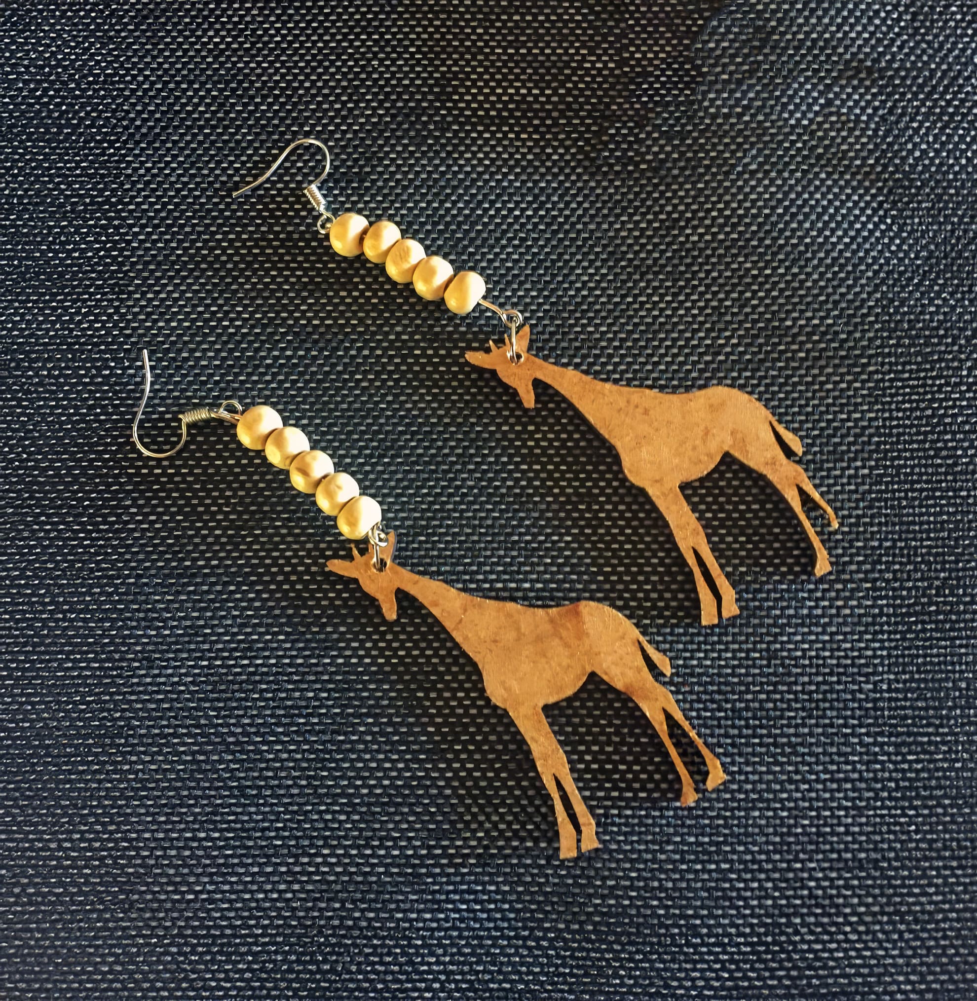 A pair of earrings with gold beads and flat, brown giraffe-shaped pendants are displayed on a textured dark fabric background. The earrings have metal hooks for wearing.