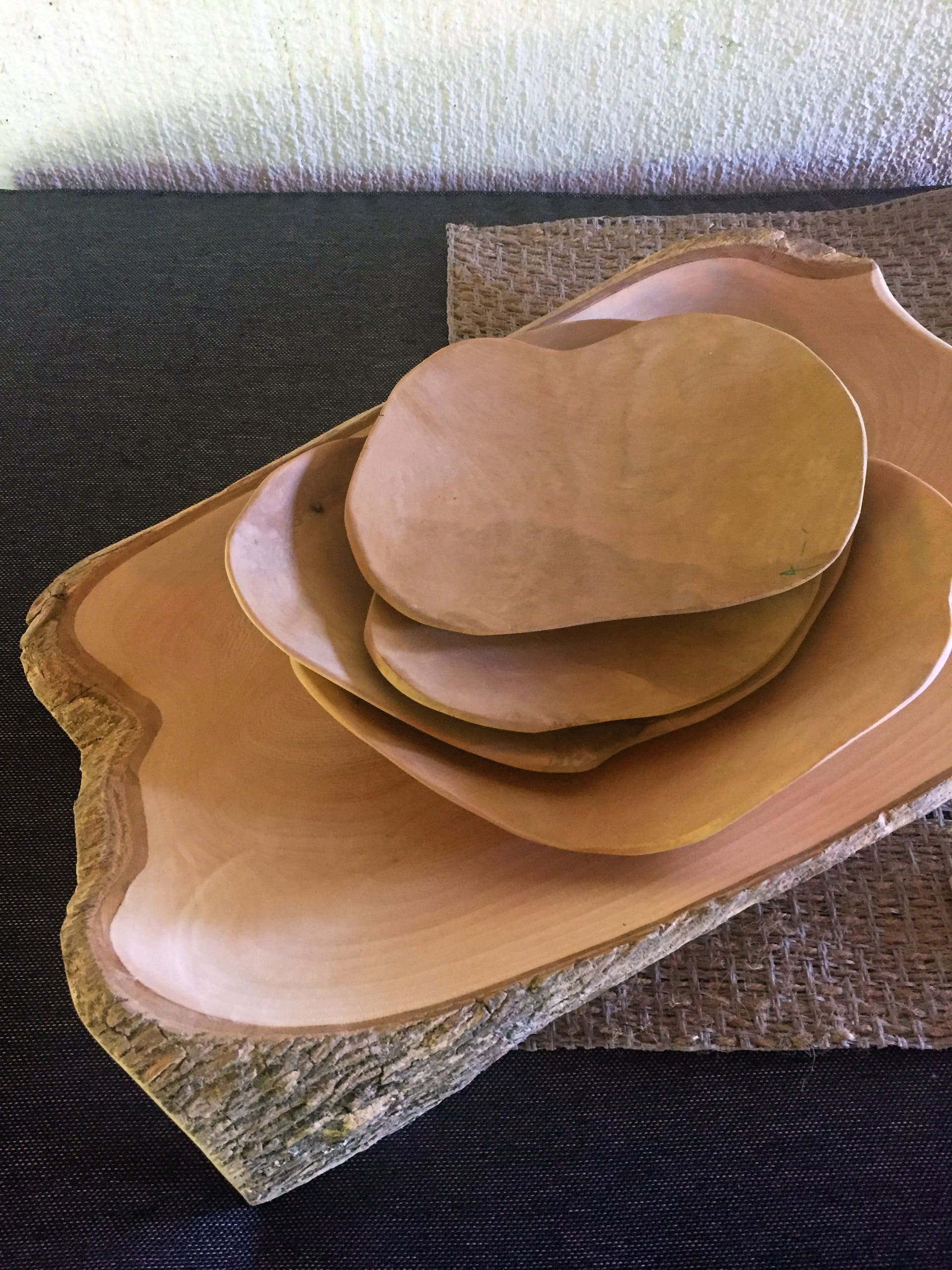 A stack of five wooden plates of varying sizes, each with irregular, organic shapes, sits on a dark fabric surface. The largest plate has a natural bark edge, and the plates rest on a woven textured mat. The background includes a pale, lightly textured wall.