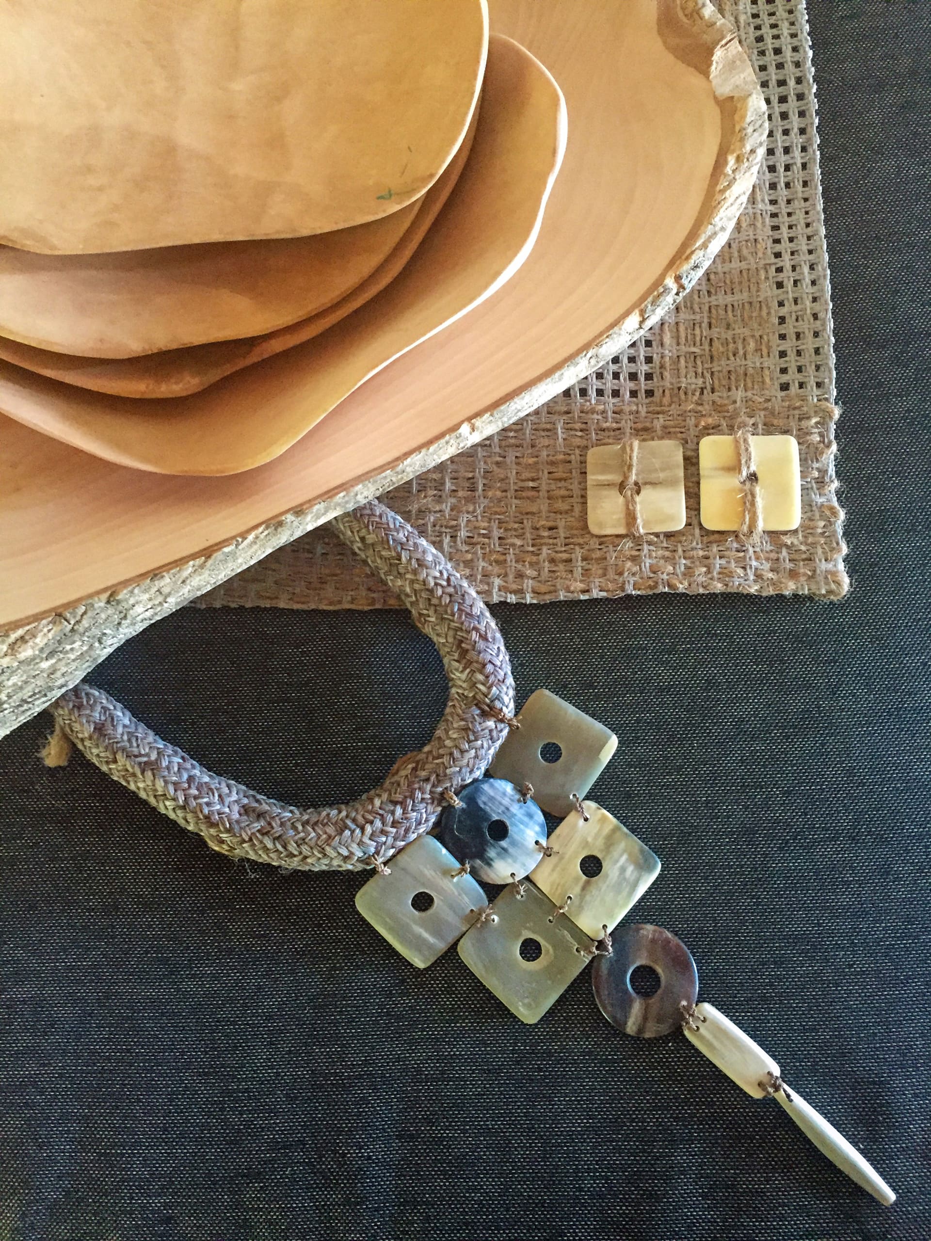Four nested wooden bowls rest on a textured brown placemat atop a dark surface. Next to the bowls, there is a woven necklace with square, round, and rectangular stone or shell pieces and a matching pair of rectangular earrings.