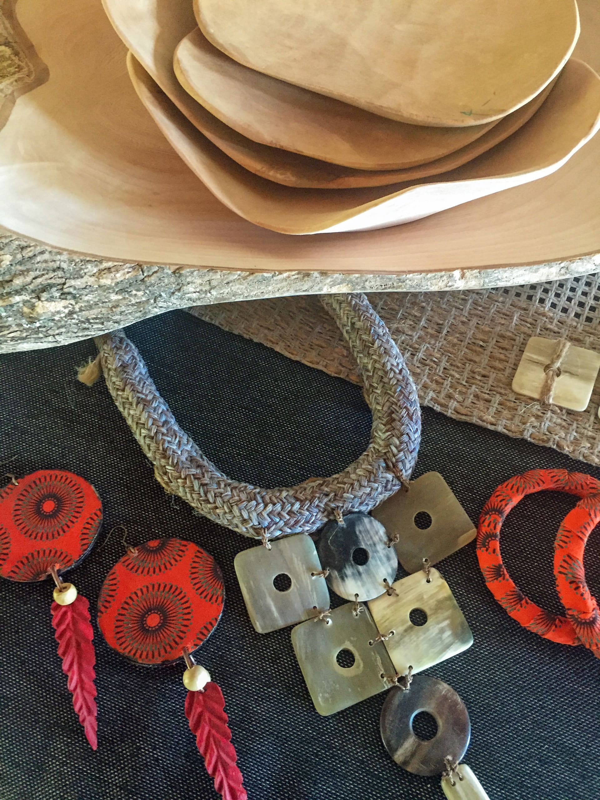 A display of hand-crafted jewellery and wooden bowls. The jewellery includes a woven necklace, two pairs of red circular earrings with feather-like pendants, and a necklace with square and round shell-like pieces. The bowls are stacked above, made of light-coloured wood.