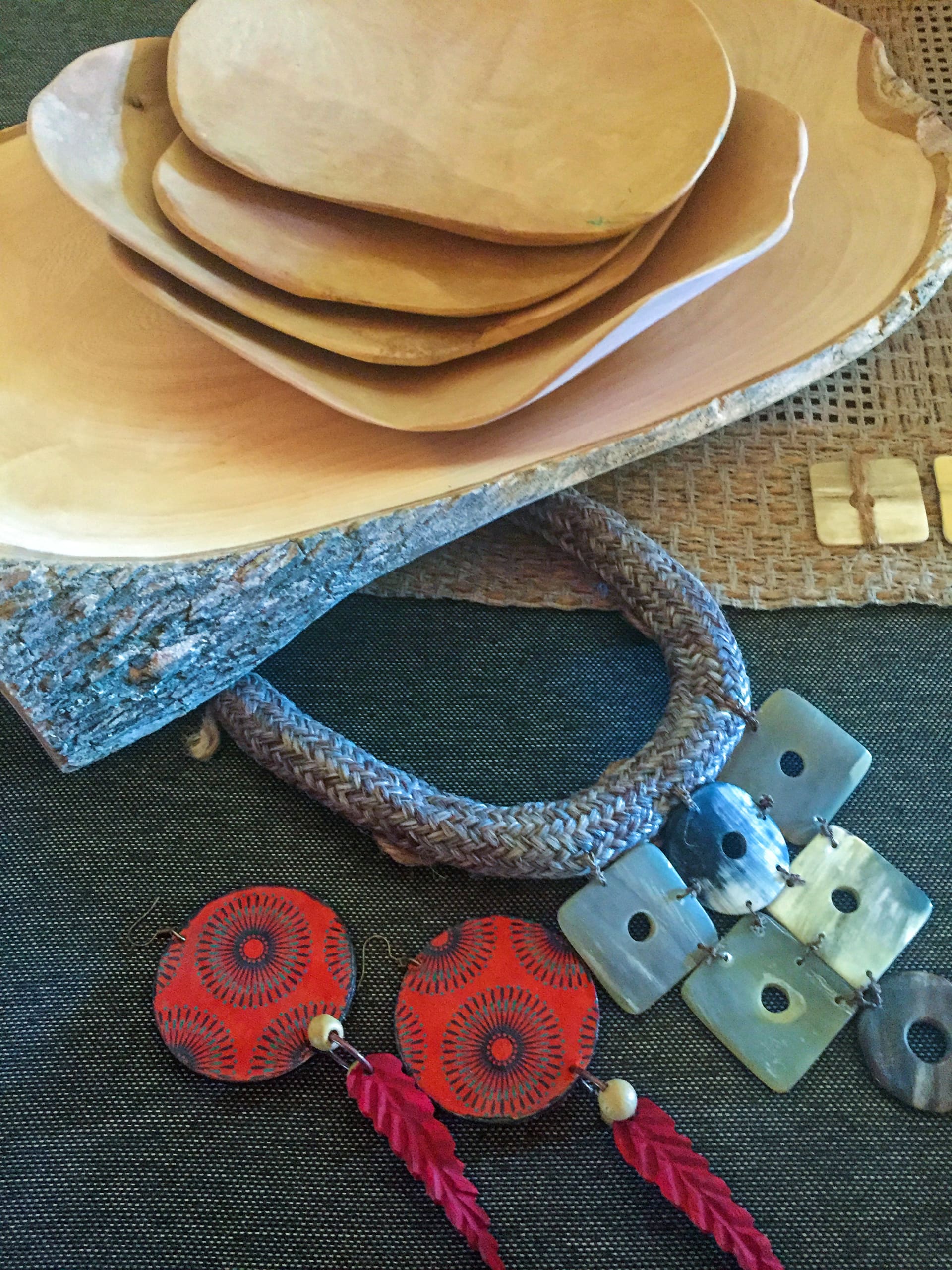 A stack of wooden plates with rustic edges sits on a woven mat. In front, there is a chunky plaited necklace with square shell pendants, and a pair of red, round earrings with feathered tassels, all placed on a dark textile background.