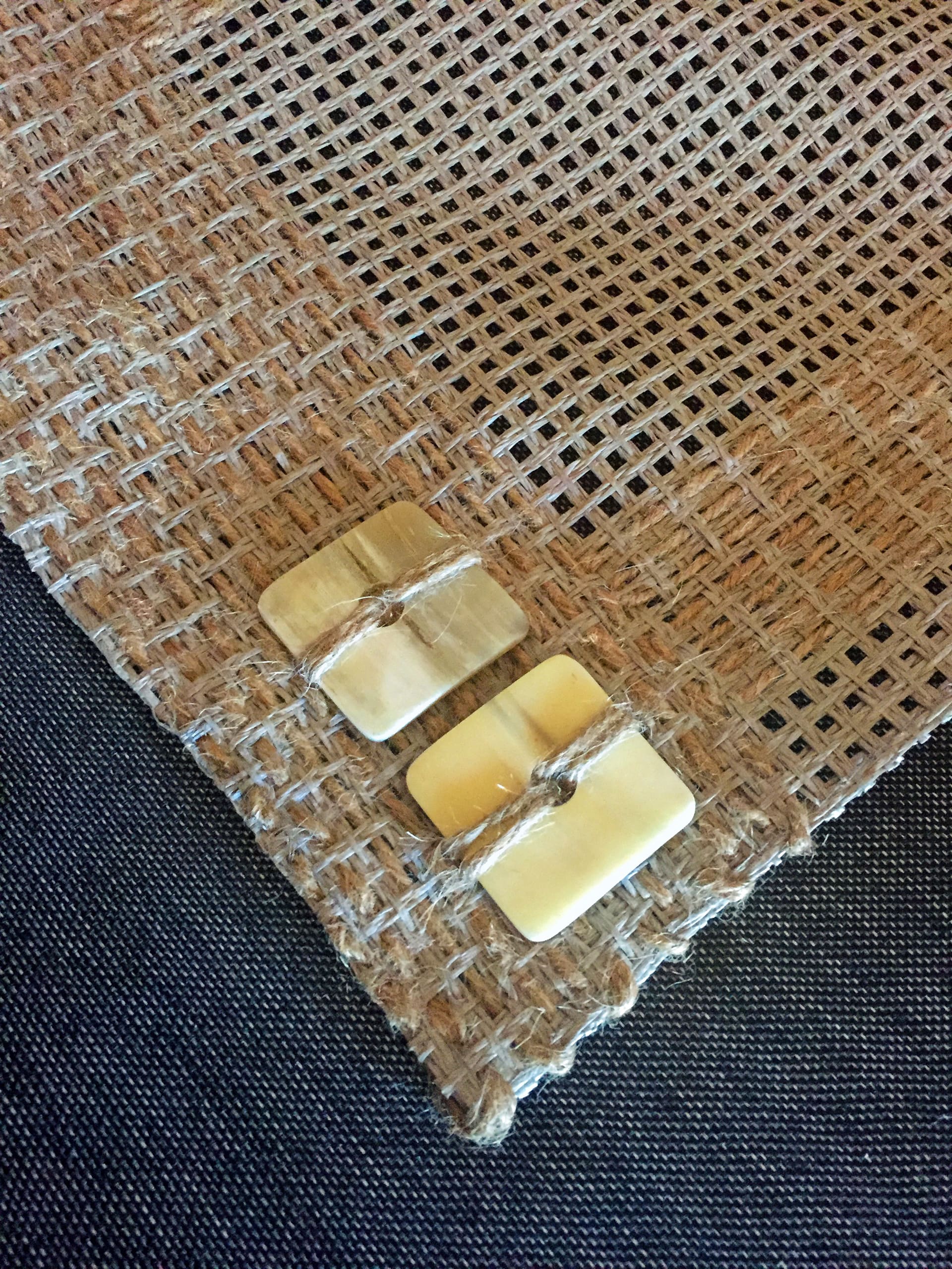 A close-up of a brown hessian fabric with two rectangular, cream-coloured buttons sewn near the edge using thick string. The background features a dark textured surface beneath the fabric.