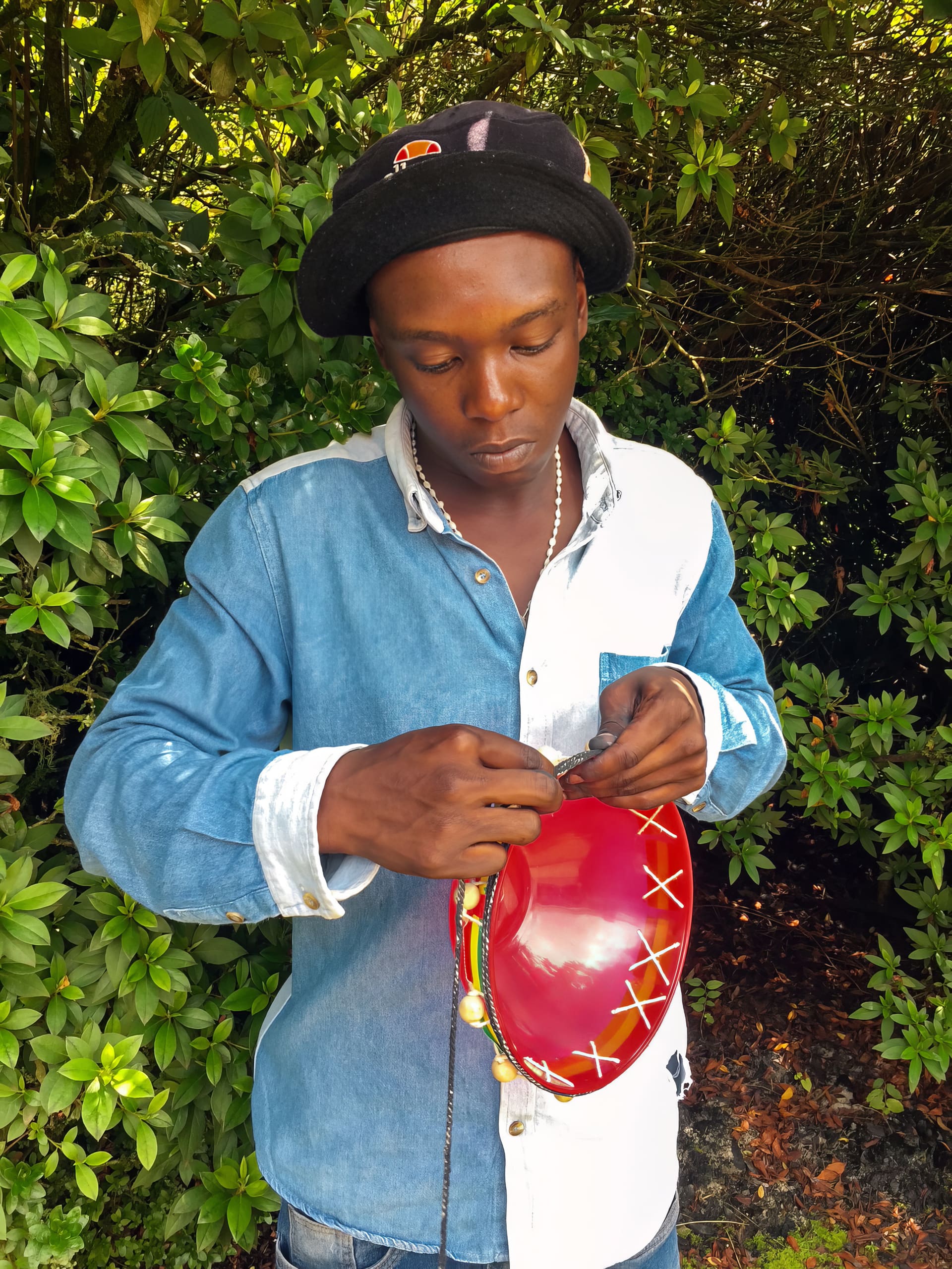 A young man in a blue and white denim shirt and black hat stands outdoors, threading string through holes on a red object, possibly a mask or decorative item, with leafy green bushes in the background. He is focused on his task.