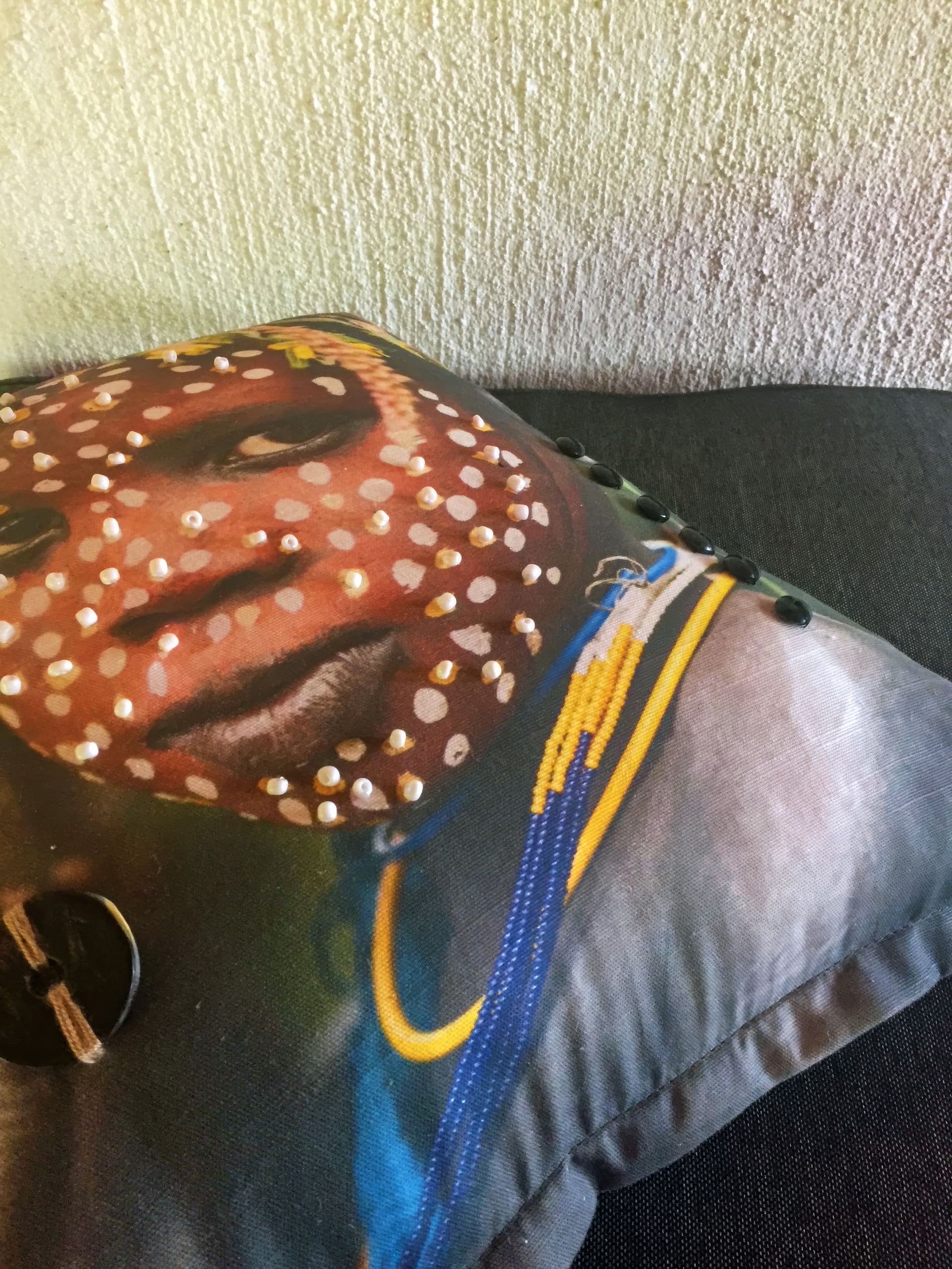 A close-up of a cushion featuring a printed image of a person with white face paint dots, wearing colourful beaded necklaces. The cushion rests on a dark sofa near a textured, off-white wall. The image has a culturally vibrant and artistic feel.
